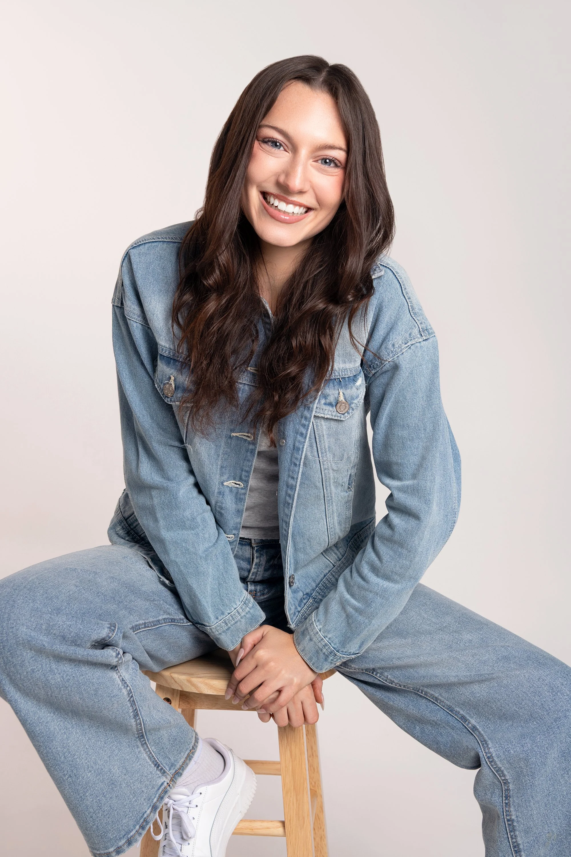 A young woman with long brown hair, smiling, wearing a denim jacket and jeans, sitting on a wooden stool against a plain light gray background.