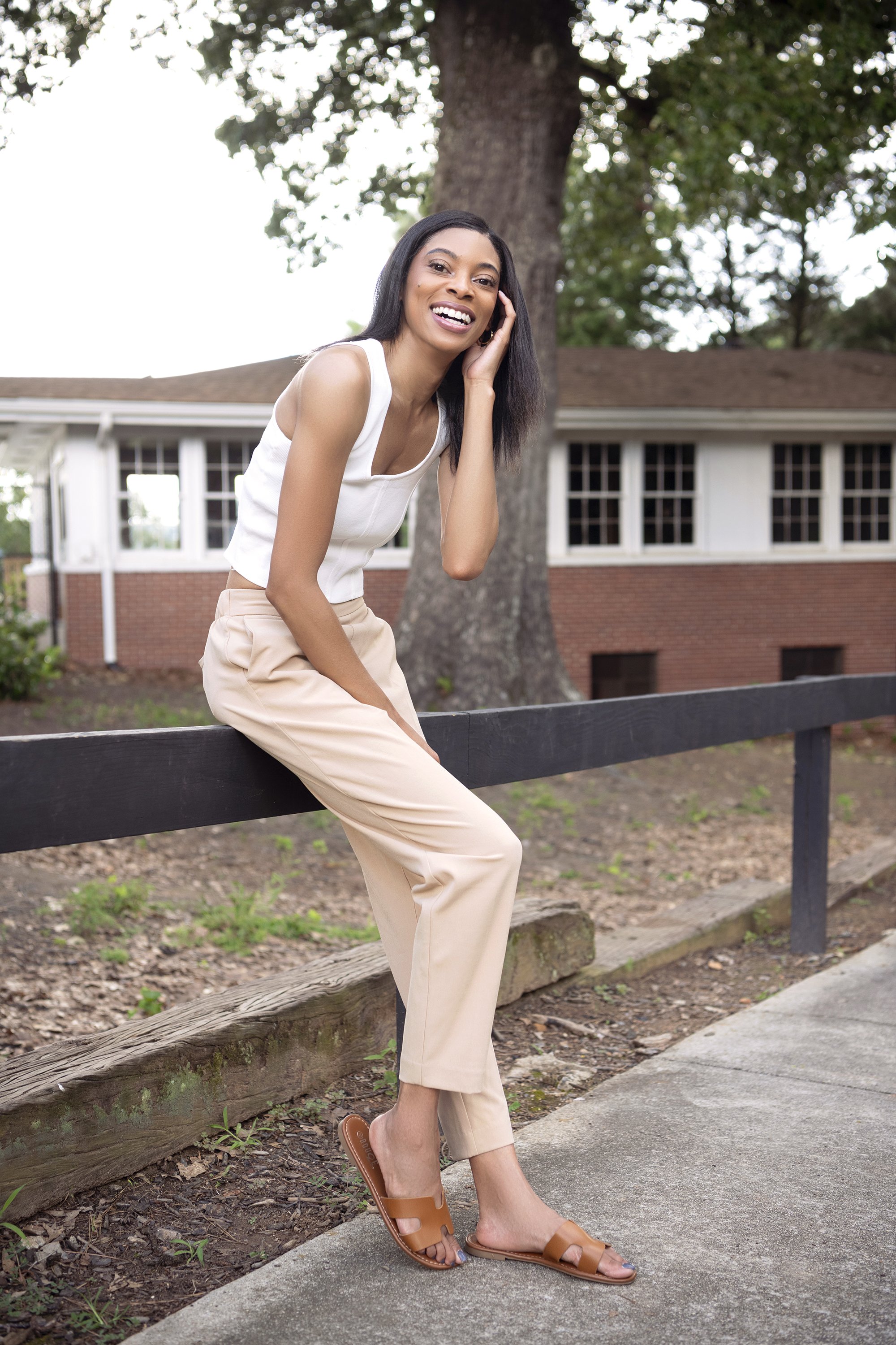 A woman with long black hair and tan skin smiling while sitting on a wooden railing outdoors near a large tree. She is wearing a white sleeveless top, beige pants, and tan sandals. There is a building with multiple windows in the background and the g