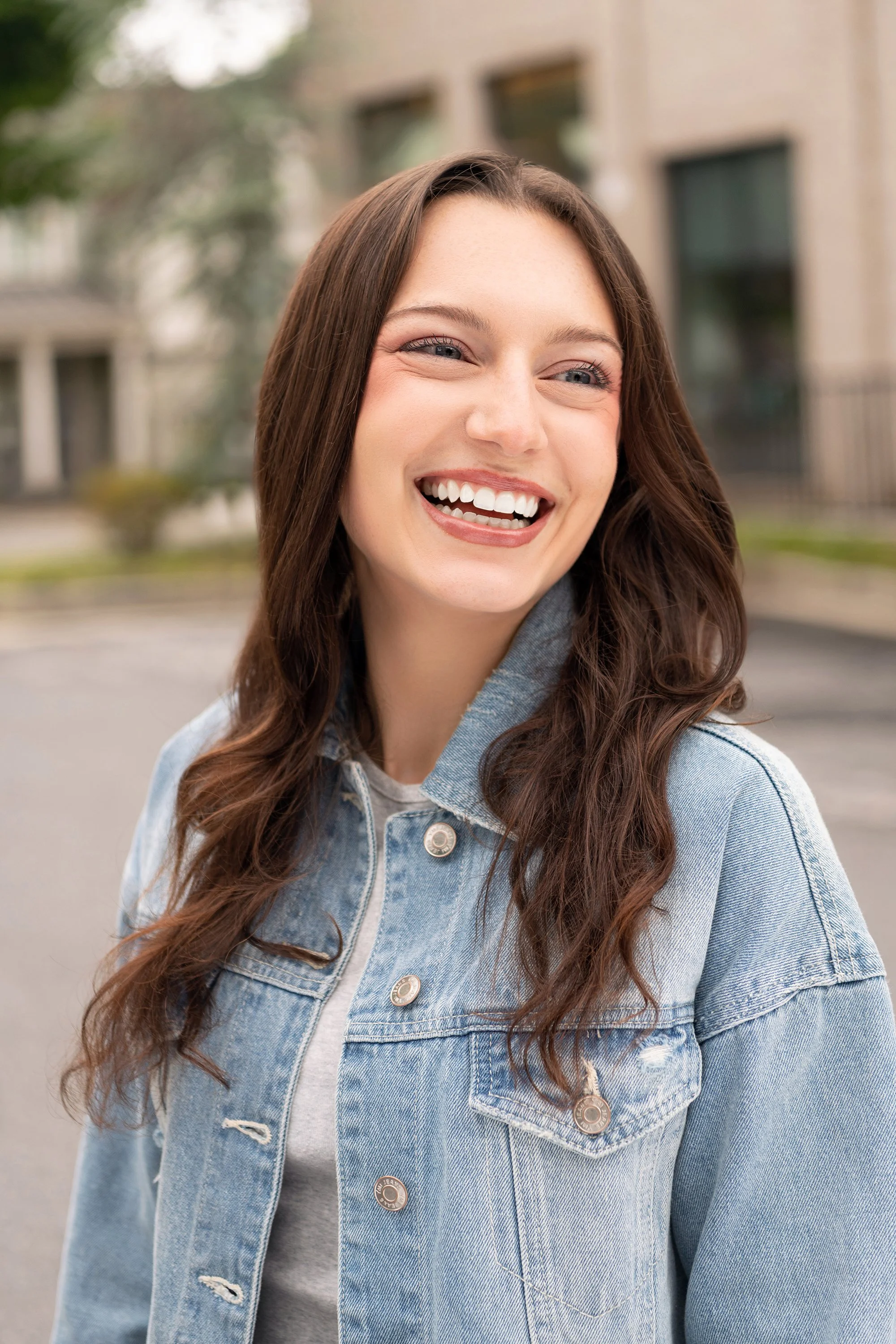 A young woman with long brown hair smiling outdoors, wearing a light denim jacket over a gray shirt, with a blurred background of buildings and trees.