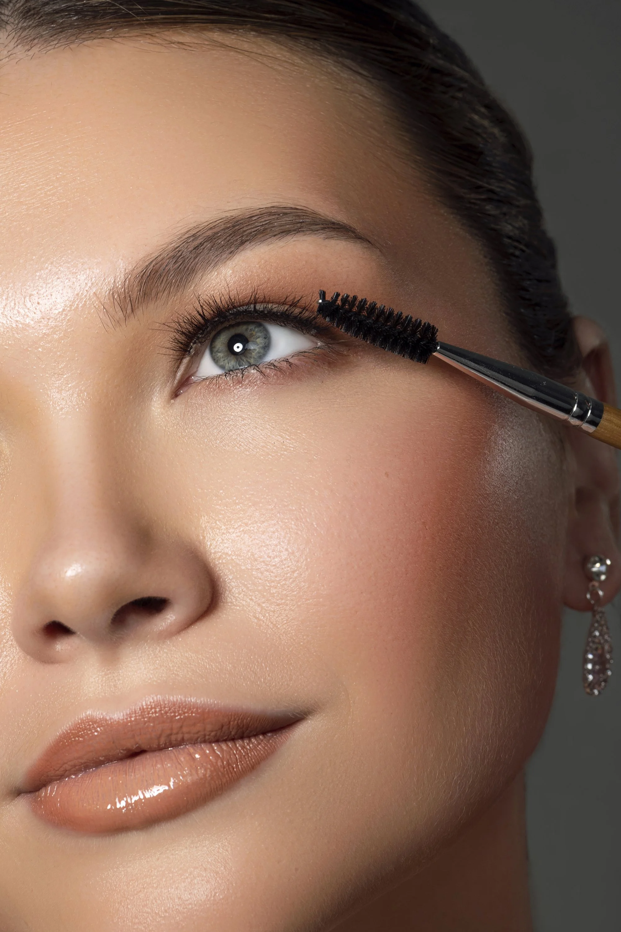 Close-up of a woman with light makeup applying mascara to her upper eyelashes.