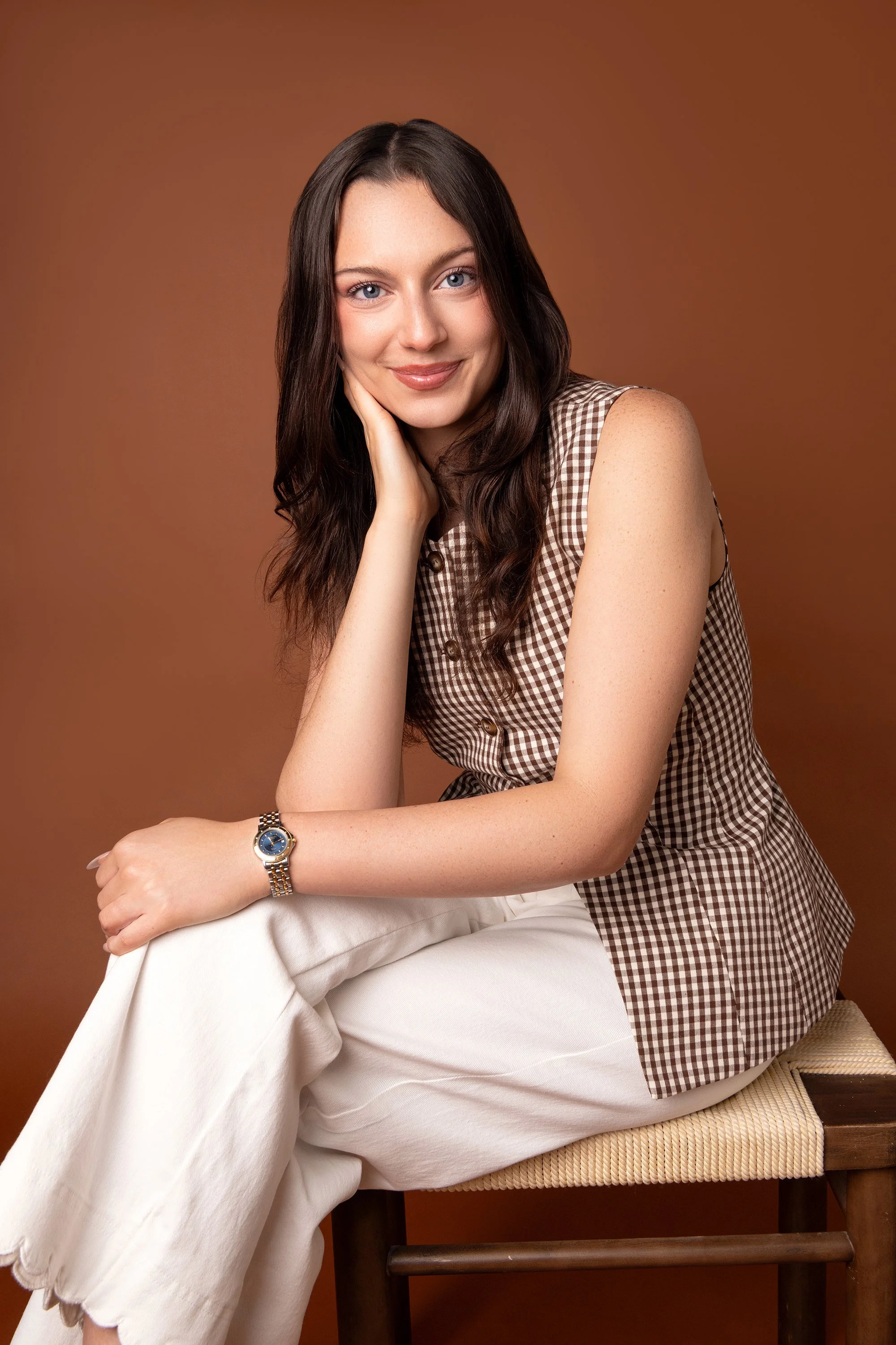 A young woman with long brown hair and blue eyes sits on a chair with a warm brown background. She is wearing a checkered sleeveless top, white pants, and a wristwatch. She rests her chin on her left hand and has a gentle smile.