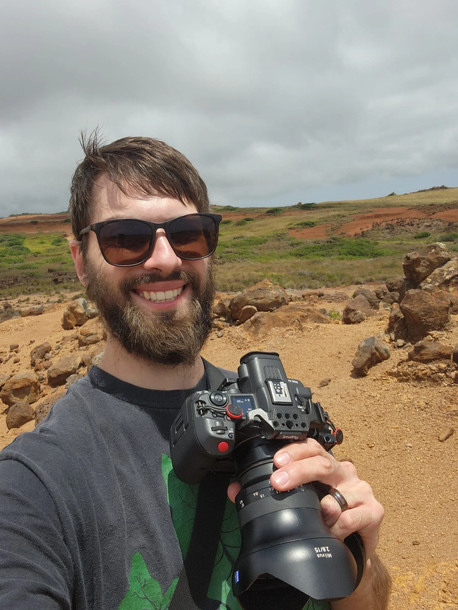 A picture of a light-skinned man with a beard and sunglasses, smiling and holding a Canon camera in an outdoor desert location.