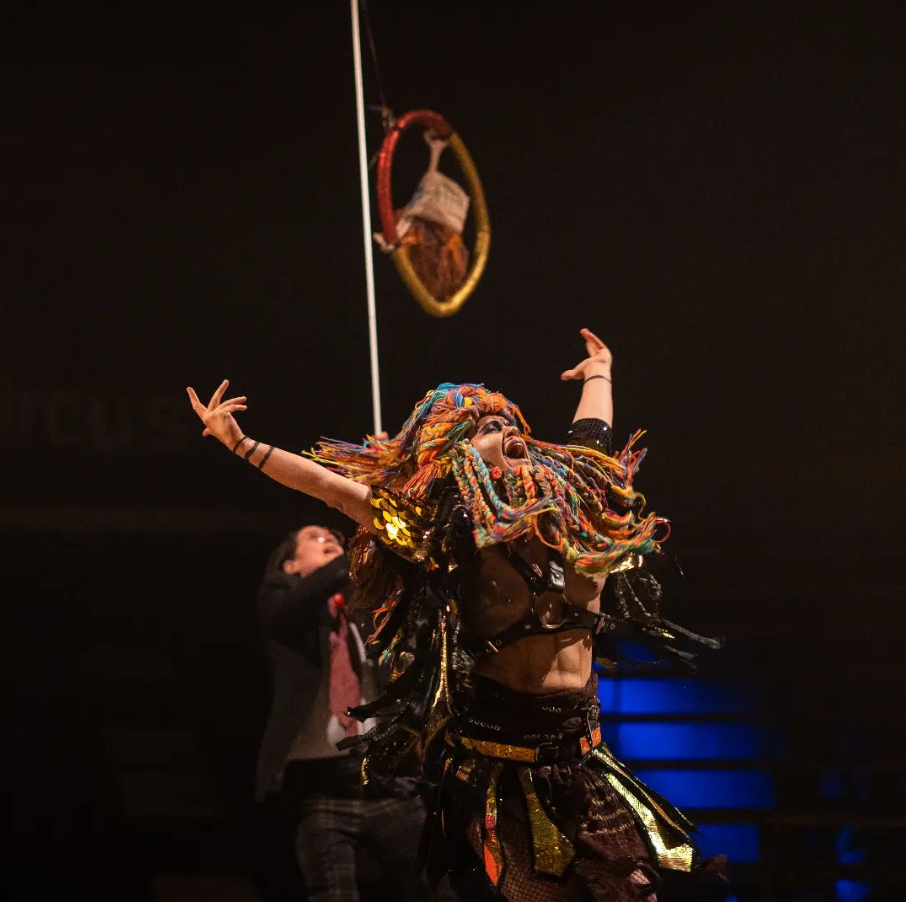 A performer with colorful dreadlocks and a vibrant costume performs on stage, with their arms raised and mouth open, while a person in the background holds a hula hoop.
