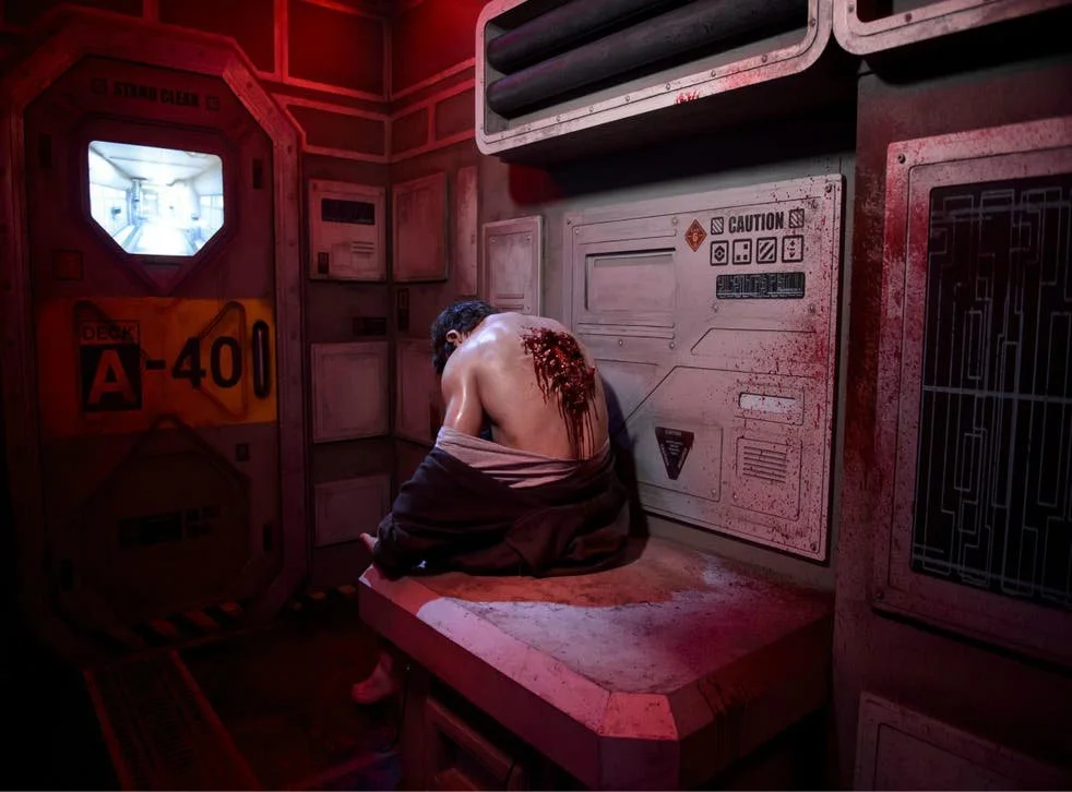 A person with long dark hair sitting on a padded bench in a dimly lit, sci-fi inspired room with red lighting. The person's back is covered in blood and they are shirtless, wearing only a dark hoodie wrapped around their waist. The room features industrial panels, warning signs, and a small window that shows a bright, white corridor outside.