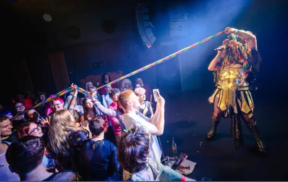 Performer with colorful hair and shiny gold outfit pulling on a rainbow-colored rope during a concert or stage performance, audience members in the background watching and recording