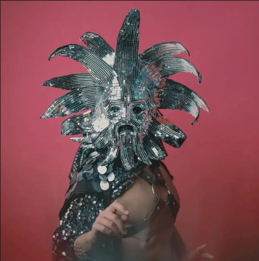 Person wearing a large, reflective, mirror-covered mask resembling a face with a wild hairdo, against a pink background.