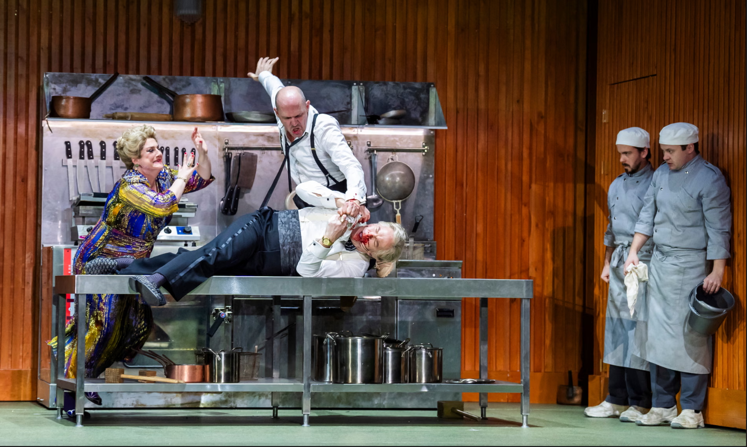 Stage scene of a theatrical play depicting a kitchen murder. A woman in a colorful dress is gesturing with her hands, a man is holding an unconscious or dead man with blood on his face, and two chefs stand at the right observing. The kitchen set has pots, pans, and utensils.