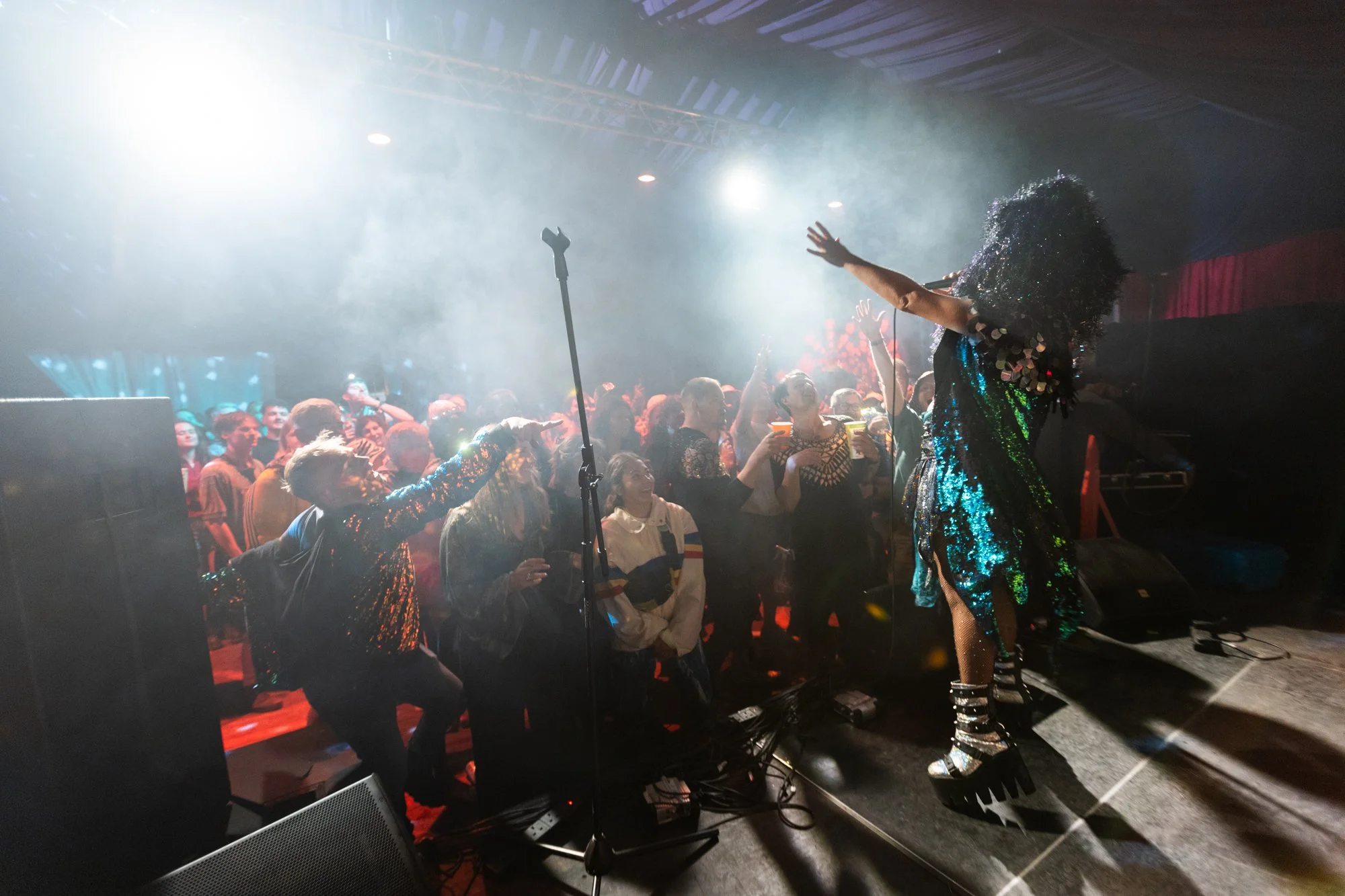 A performer with curly hair and a shiny, colorful dress dancing on stage in front of a lively crowd at a concert or nightclub, with fans reaching out towards her.