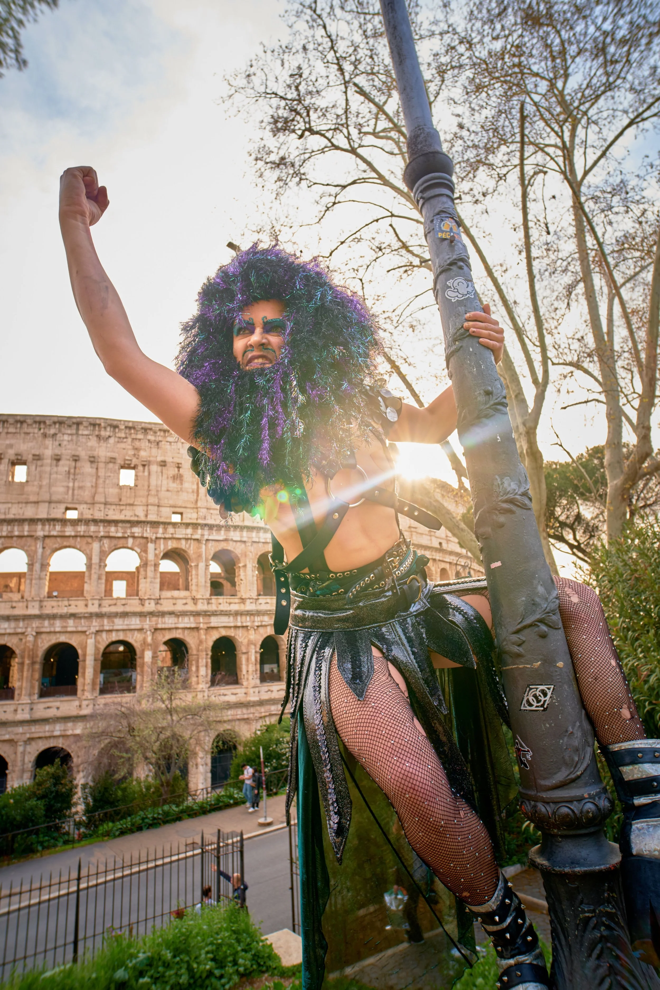Person wearing a large, colorful curly wig, face paint, and a revealing artistic costume, holding onto a lamp post in front of the Roman Colosseum.