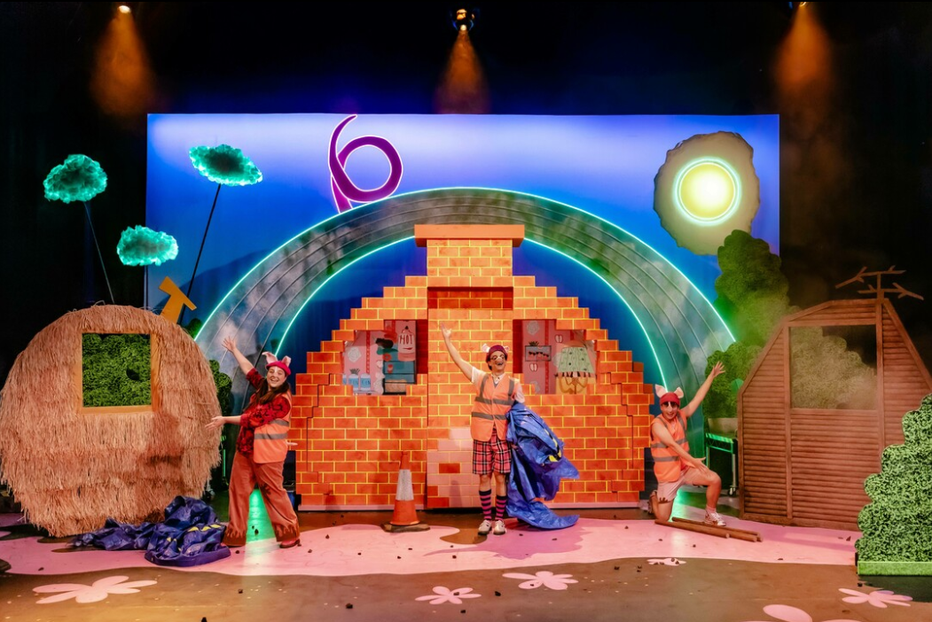 Four performers dressed as construction workers on a colorful stage with a whimsical set. The set includes a brick wall with openings, a thatched hut, a wooden shed, and painted sun and cloud backdrop with a rainbow. The performers are smiling and waving, with one holding a blue tarp, and two wearing pink hats with pig ears.