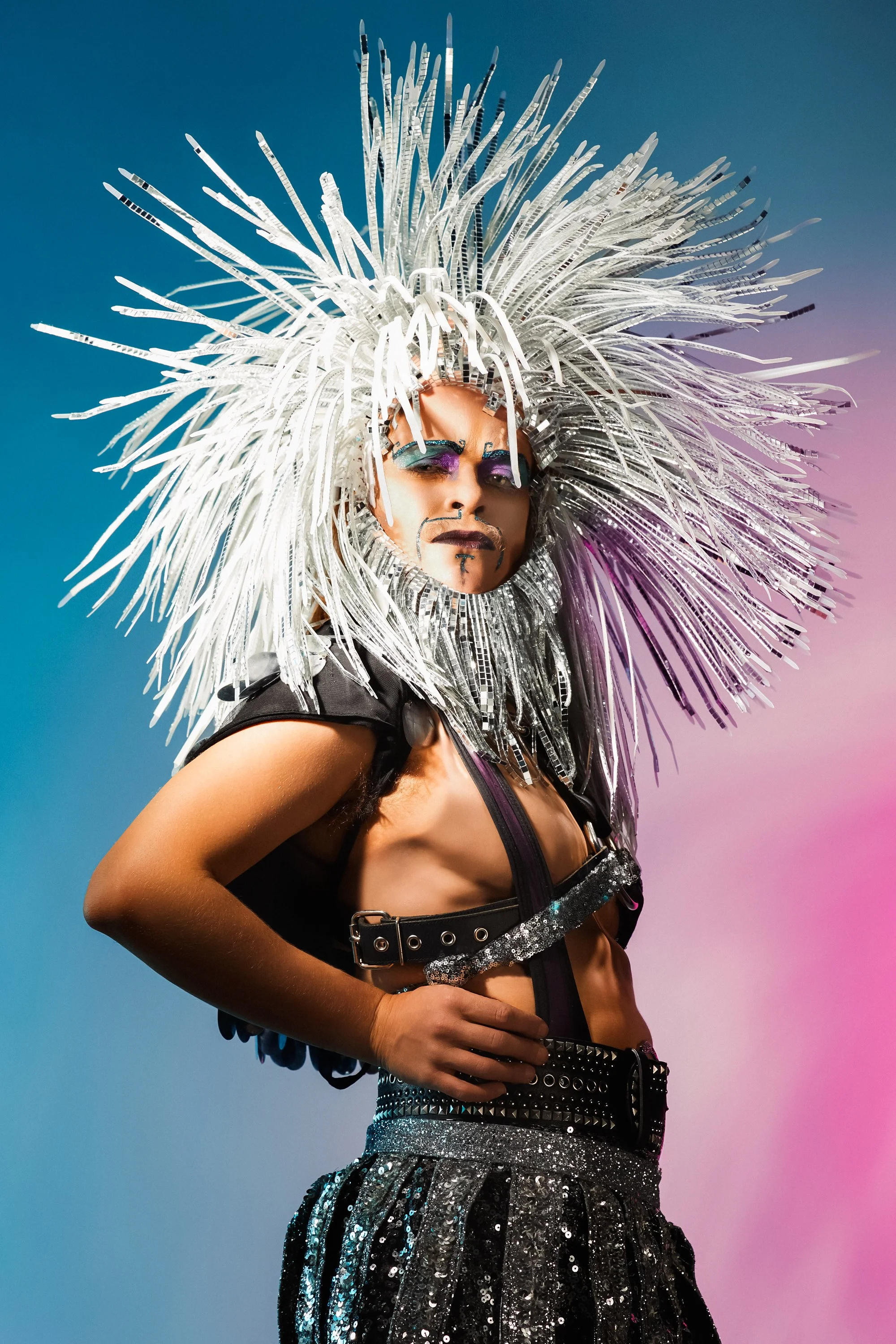Person dressed in a silver, metallic costume with an elaborate, spiky headdress, makeup, and colorful eye makeup standing against a gradient background.