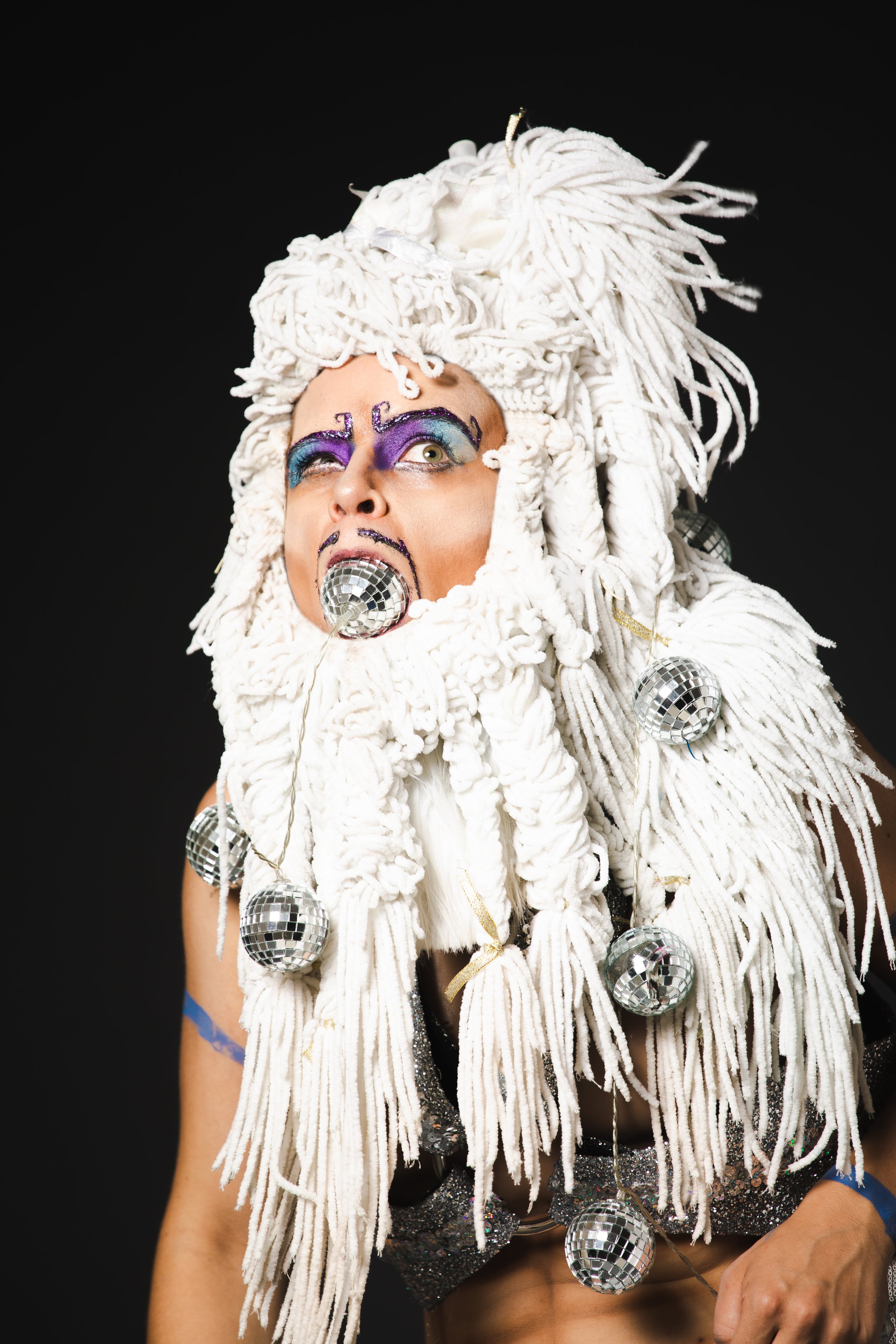Person dressed in a creative costume resembling a white yarn and disco balls, with vibrant makeup and a black background.