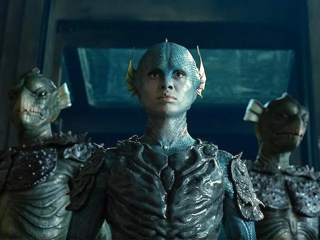 A humanoid alien with blue skin and fin-like structures on its head, dressed in black armor, standing inside a spaceship with two fish-like creatures with fins and armored bodies flanking it.