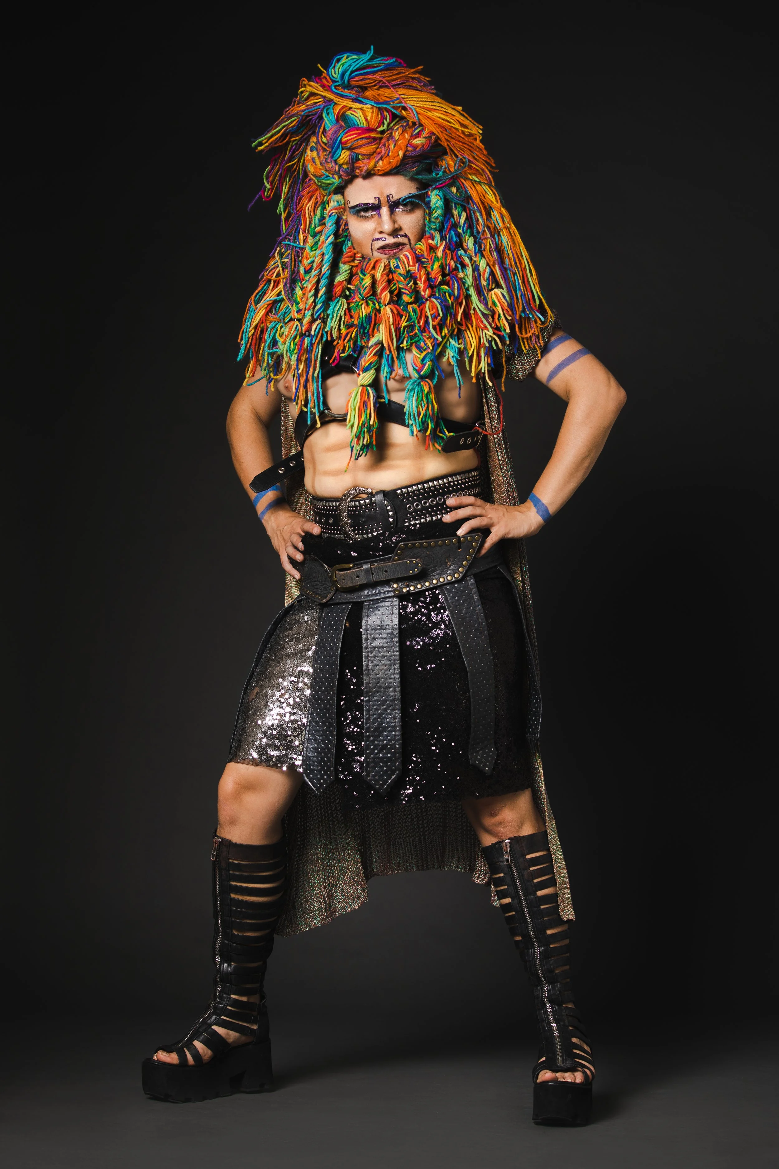 A person with a rainbow-colored dreadlock wig, heavy makeup, and bold clothing in a studio setting with a dark background.