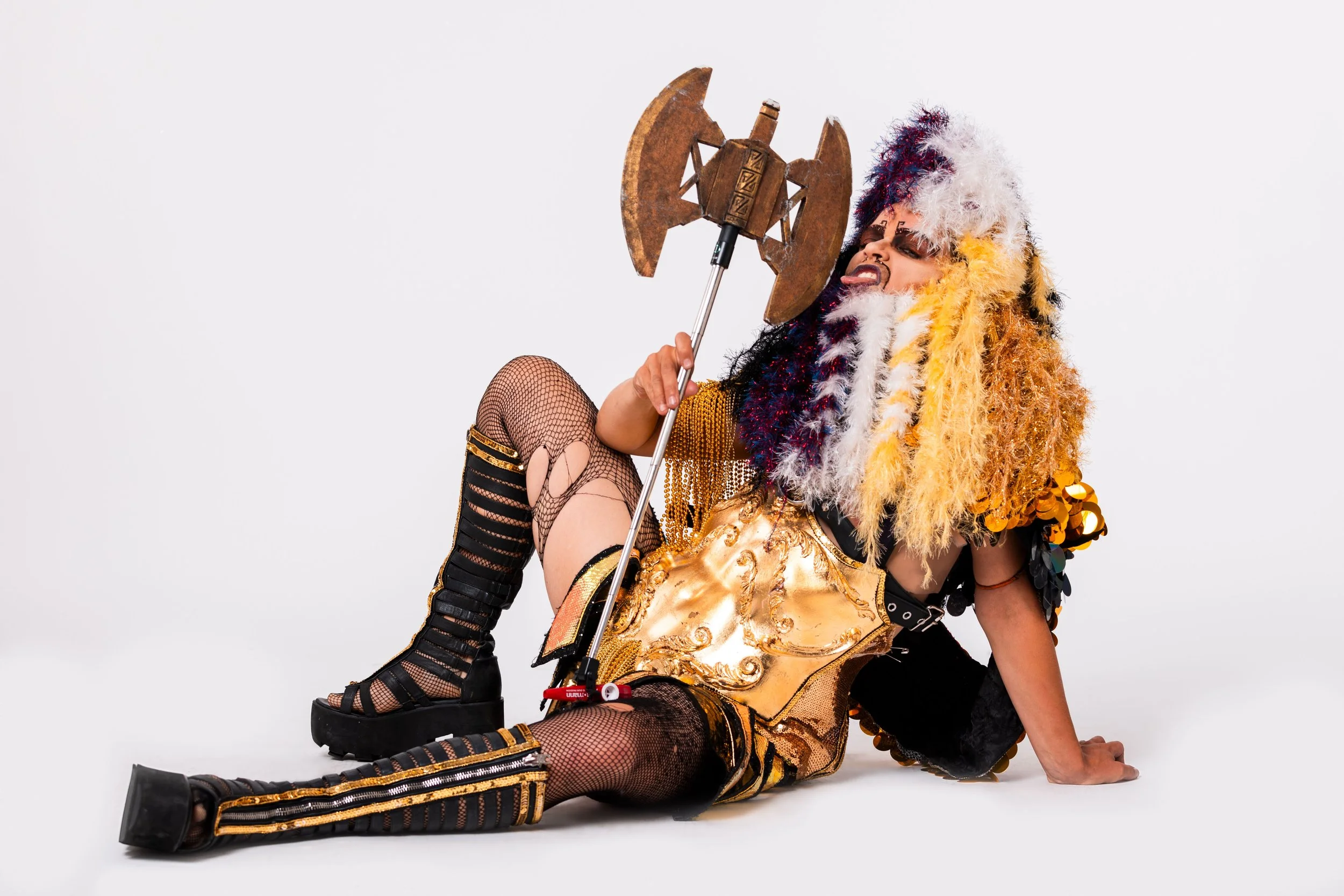 Person in elaborate costume with gold armor, colorful wig, fishnet stockings, and platform shoes sitting on the floor, holding a decorative axe.