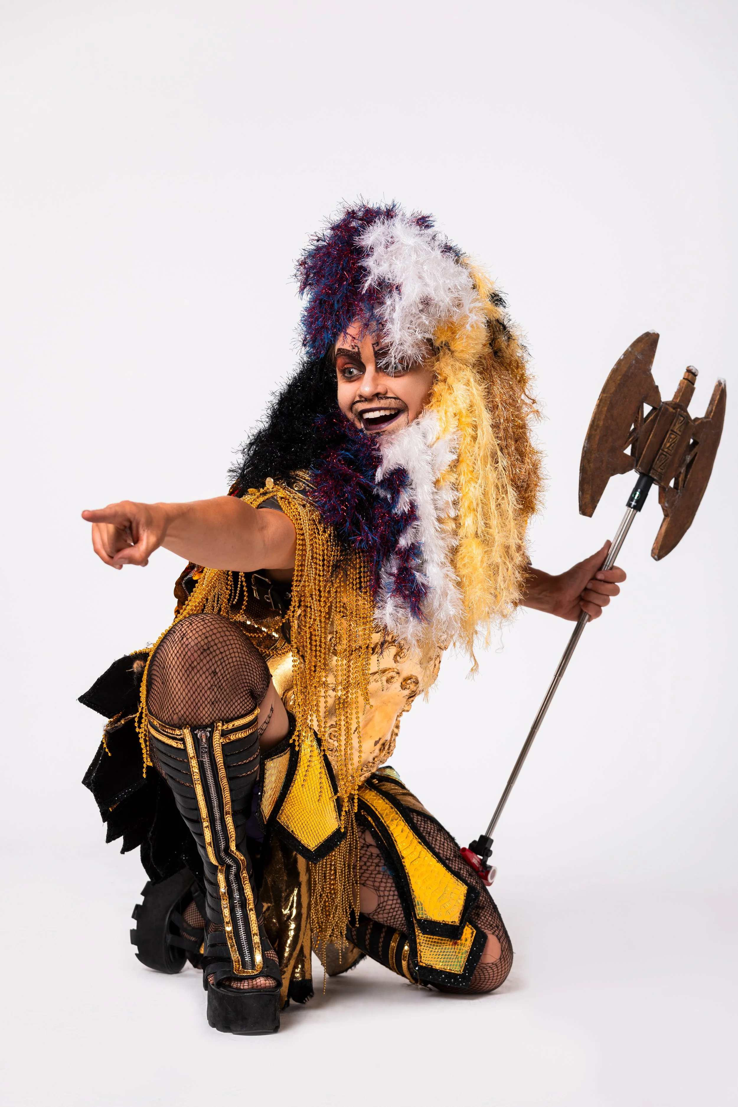 Person in a colorful costume with feathers and chains, holding a prop axe, smiling and pointing forward.