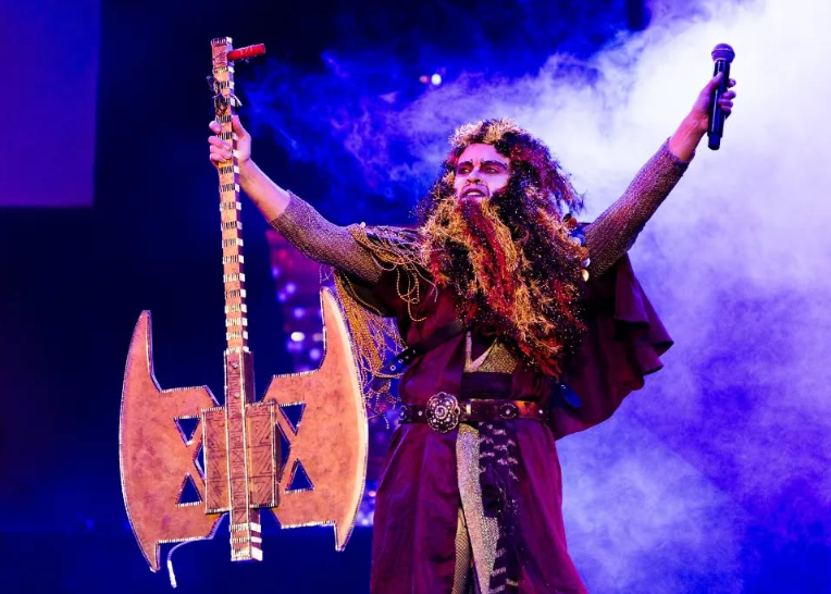 A person dressed as a fantasy warrior on stage, holding a large axe prop in one hand and a microphone in the other, with dramatic lighting and fog effects.