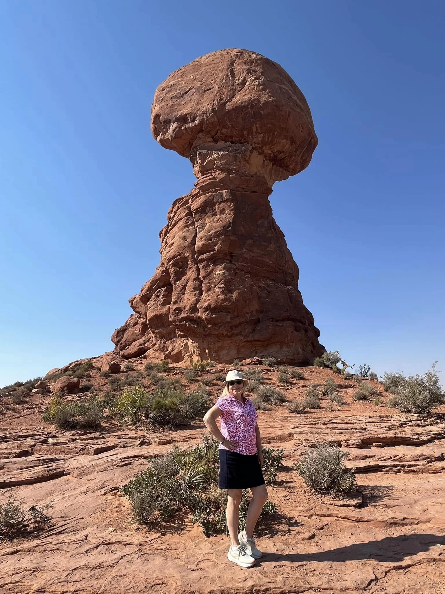 A woman wearing a pink patterned shirt, black skirt, white hat, and sunglasses stands in front of a large red rock formation in a desert landscape under a clear blue sky.