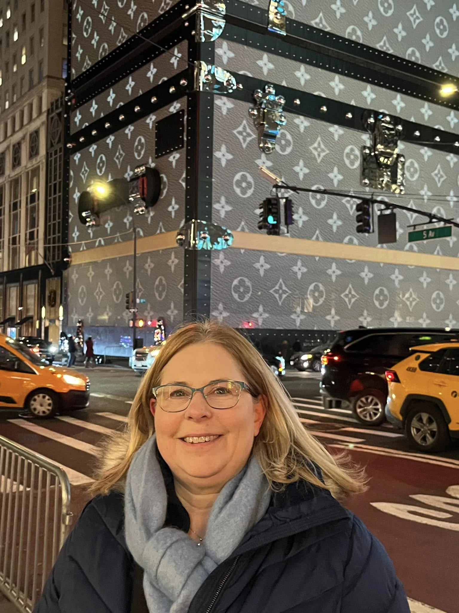 A smiling woman with glasses stands on a city street at night, in front of a building that has a large Louis Vuitton patterned decoration projected onto it, with traffic and pedestrians in the background.