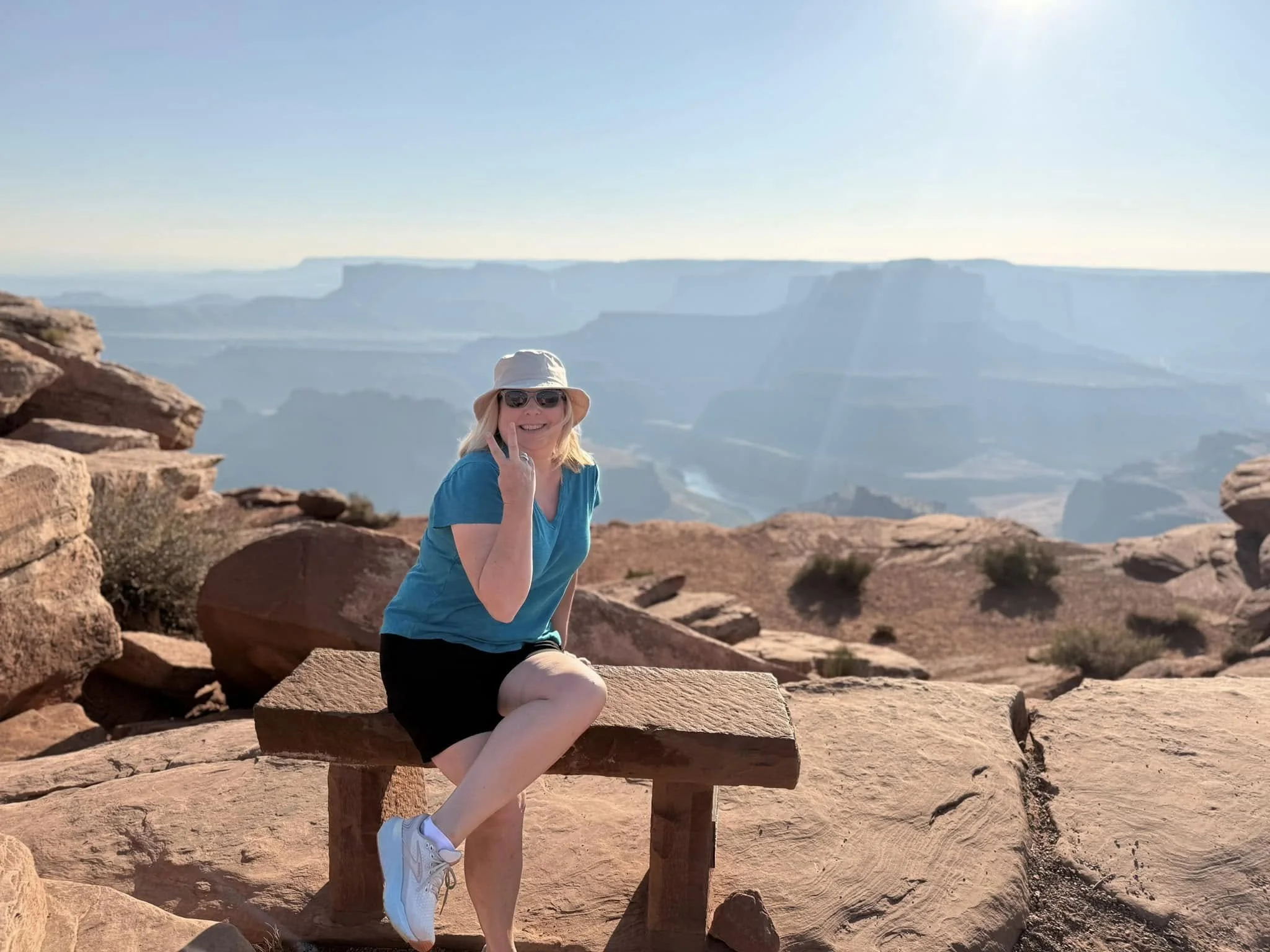 Woman sitting on a wooden bench at the Grand Canyon, smiling, with a sunhat, sunglasses, blue shirt, black shorts, and white sneakers, with the canyon in the background.