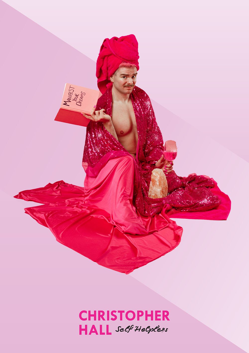 Christopher Hall as Jeff Hopeless sitting on a pink floor draped with pink fabric, wearing a large pink towel on his head, a sequined pink robe, holding a pink cocktail and salt lamp, with a pink notebook titled "Manifest Your Dreams" in the backgrou