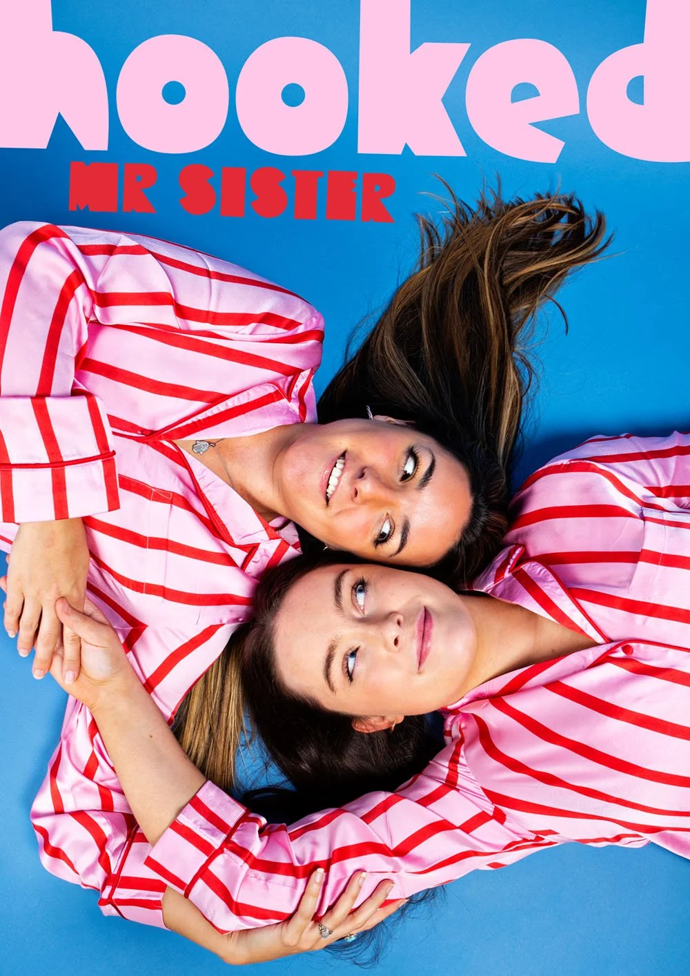 Two women with long hair lie on a blue background, wearing matching pink and red striped pajamas, smiling and looking at each other. There is pink and red text on the background that says "hooked Mr Sister."