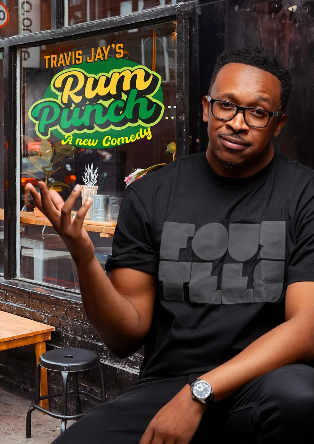 A man with glasses and a black t-shirt sitting inside a cafe, gesturing with his hand, in front of a colorful sign that reads 'Travis Jay's Rum Punch: A new Comedy.'