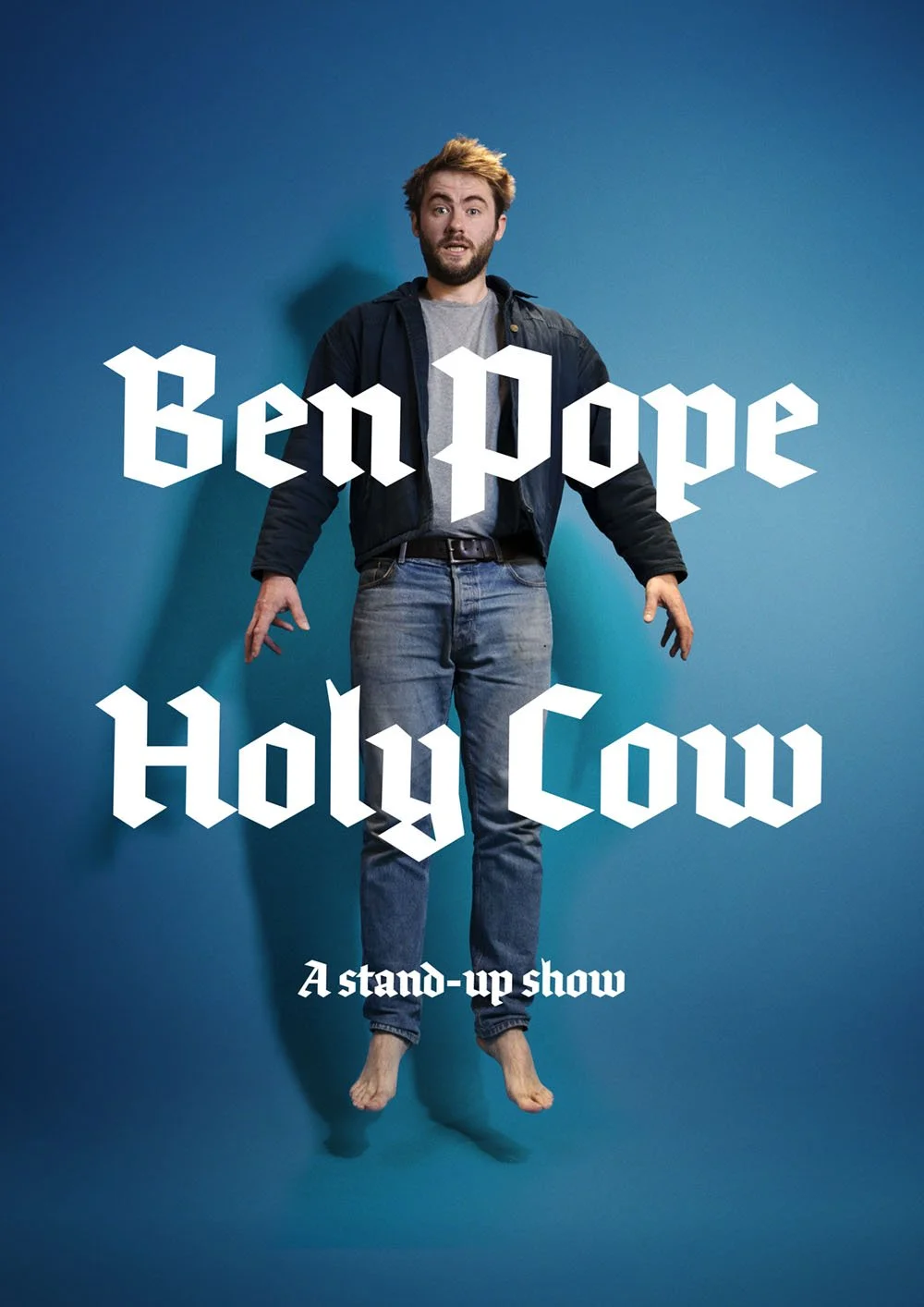 Poster for Ben Pope and Holly Cow stand-up show featuring a man with messy hair, beard, gray t-shirt, jeans, and a jacket, floating barefoot against a blue background, with the title text.