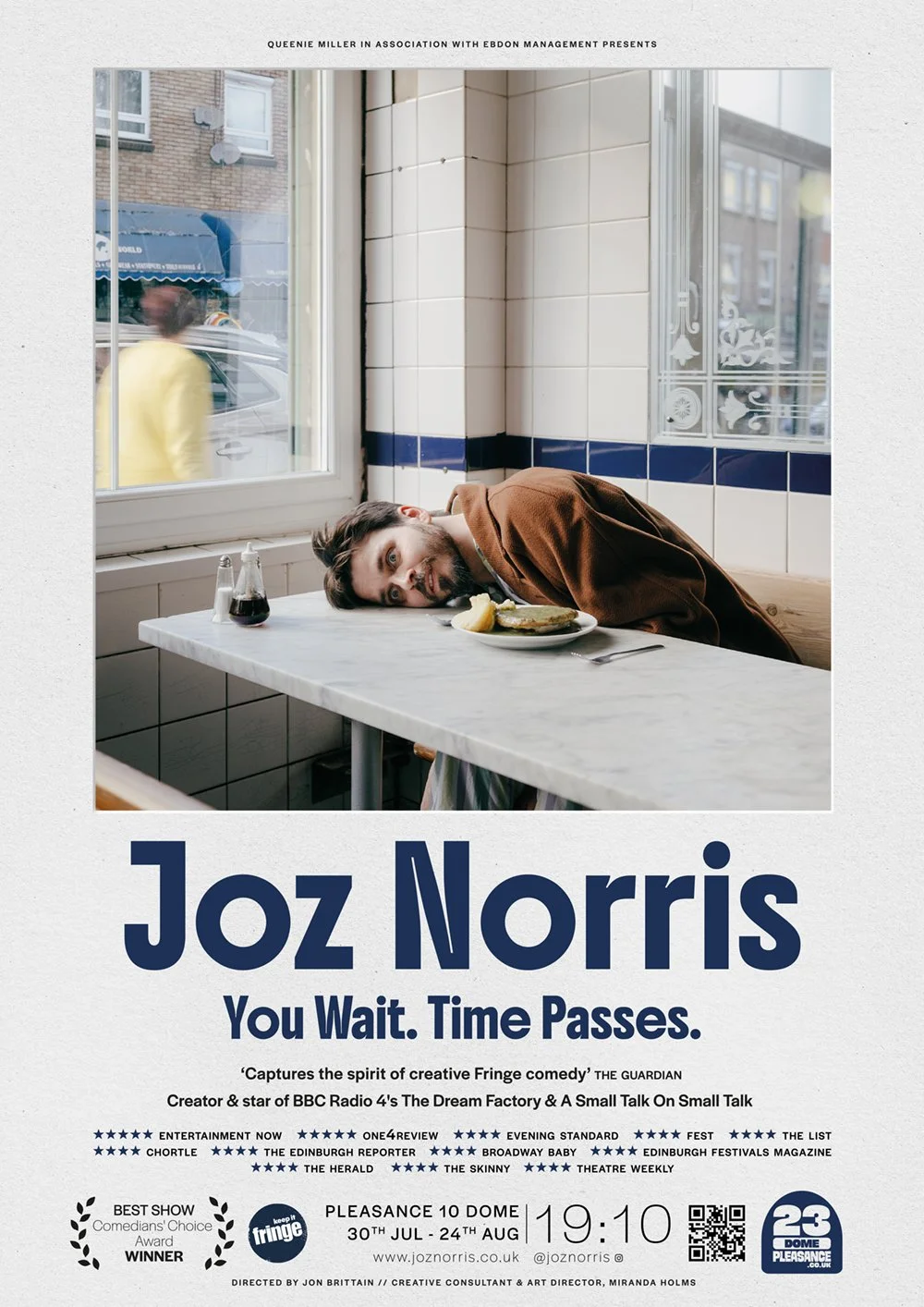 A man with dark hair and a beard, leaning on a table in a restaurant, looking at the camera with a playful expression, with a plate of food and condiments on the table behind him.