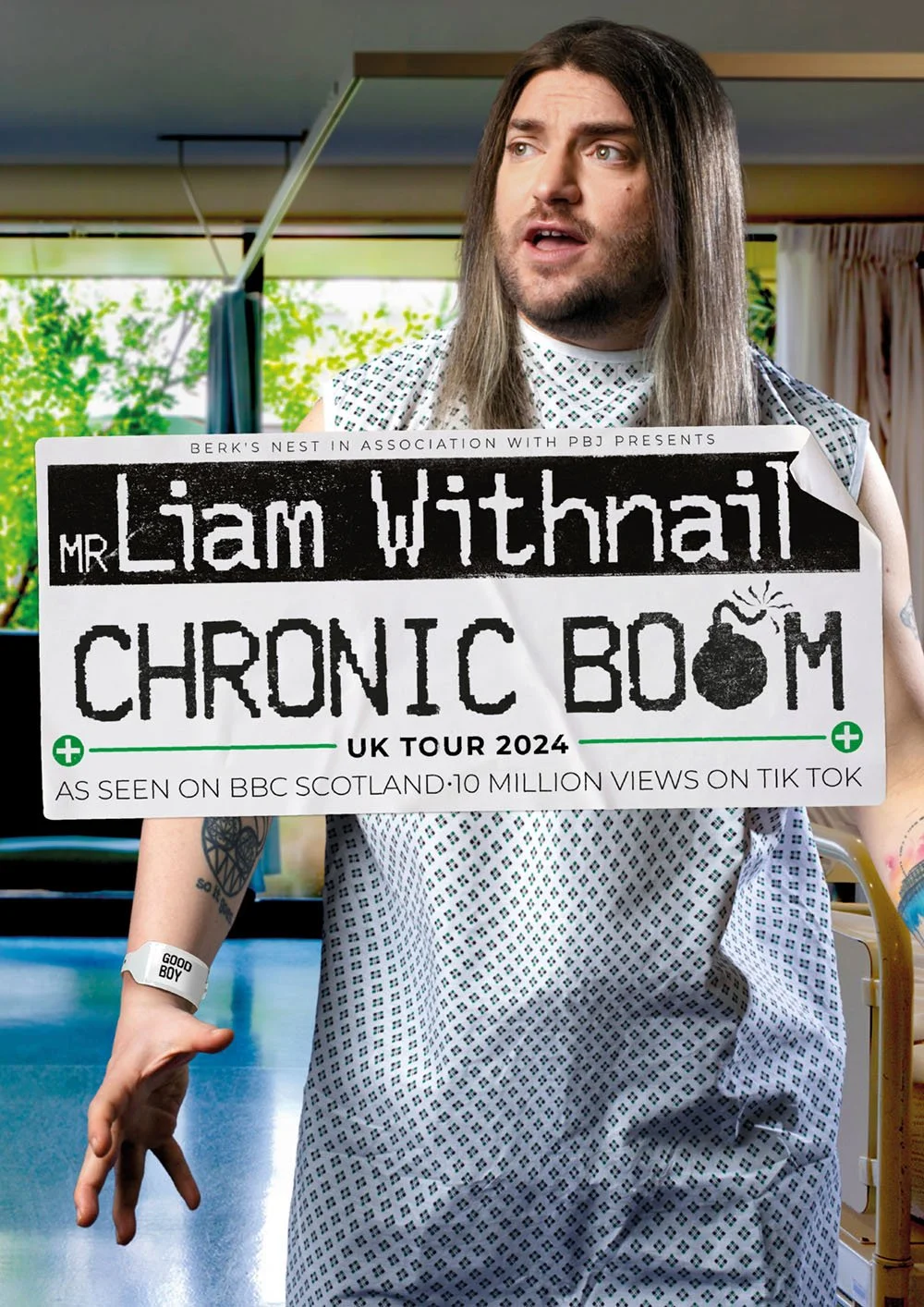 Man with long dark hair and tattoos on his arm holding a promotional poster for Liam Withnail's 'Chronic Boom' UK tour 2024, wearing a hospital gown, standing in a room with windows and curtains.