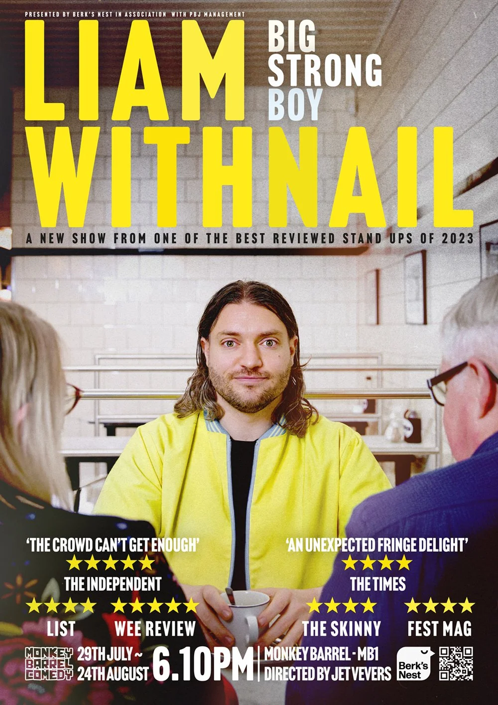 Poster for the comedy show 'Liam Withnall: Big Strong Boy' featuring a man with shoulder-length dark hair and a beard, wearing a yellow jacket, sitting at a table with two audience members.