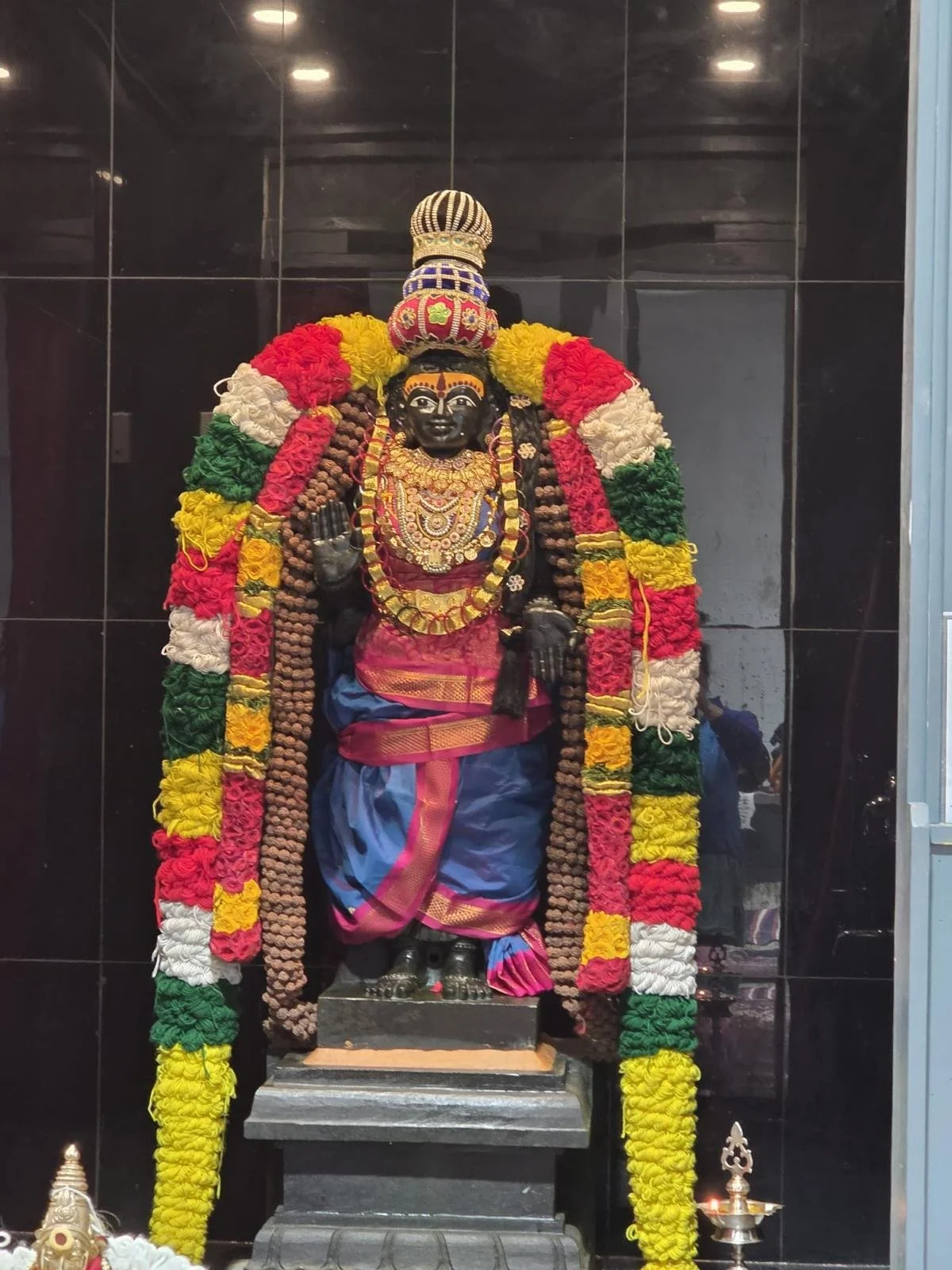 Decorative Hindu deity sculpture dressed in yellow and blue silk saree, adorned with jewelry, flower garlands, and traditional ornaments, with two smaller deity statues in colorful silk garments at the base.