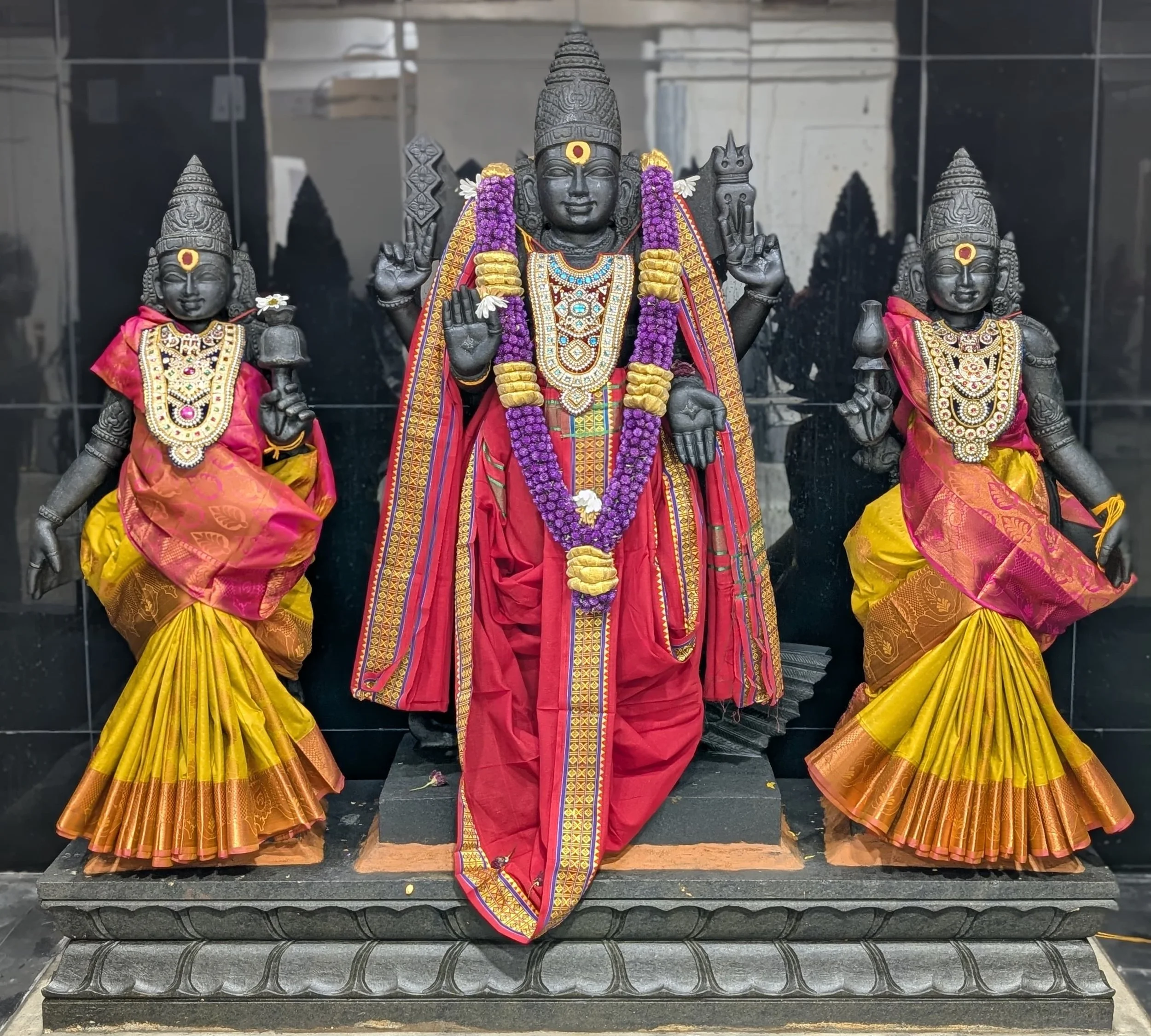 Hindu temple statues of a central black stone deity adorned with a purple flower garland and colorful cloth, flanked by two smaller statues dressed in pink and yellow garments with jewelry and floral decorations.