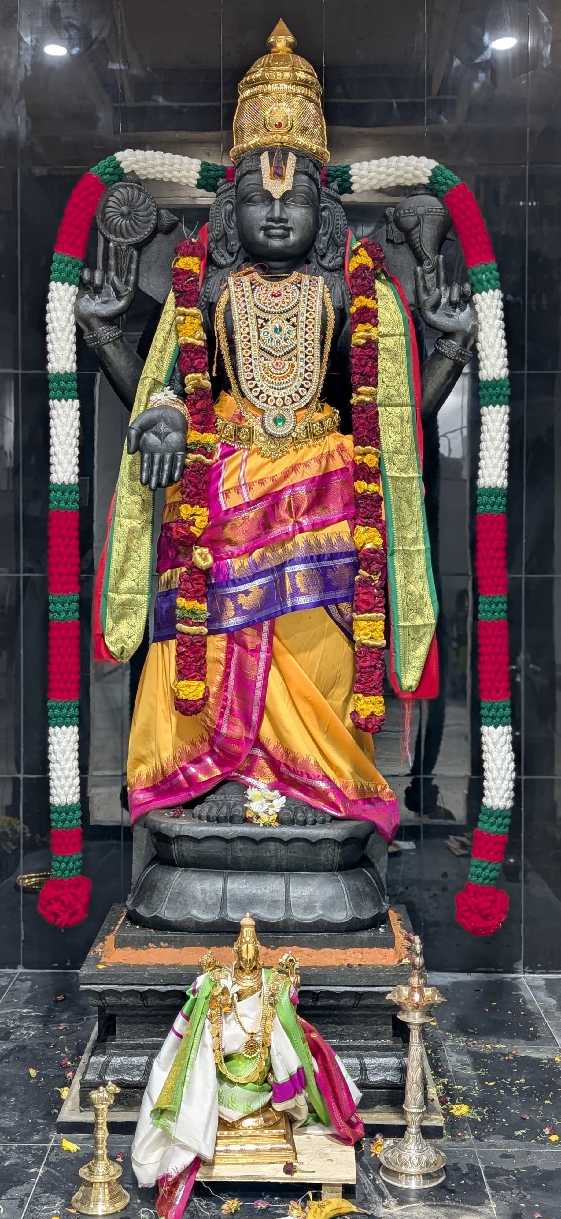 Hindu deity statue dressed in colorful silk clothing and adorned with intricate jewelry, with a garland of flowers, in a temple setting.