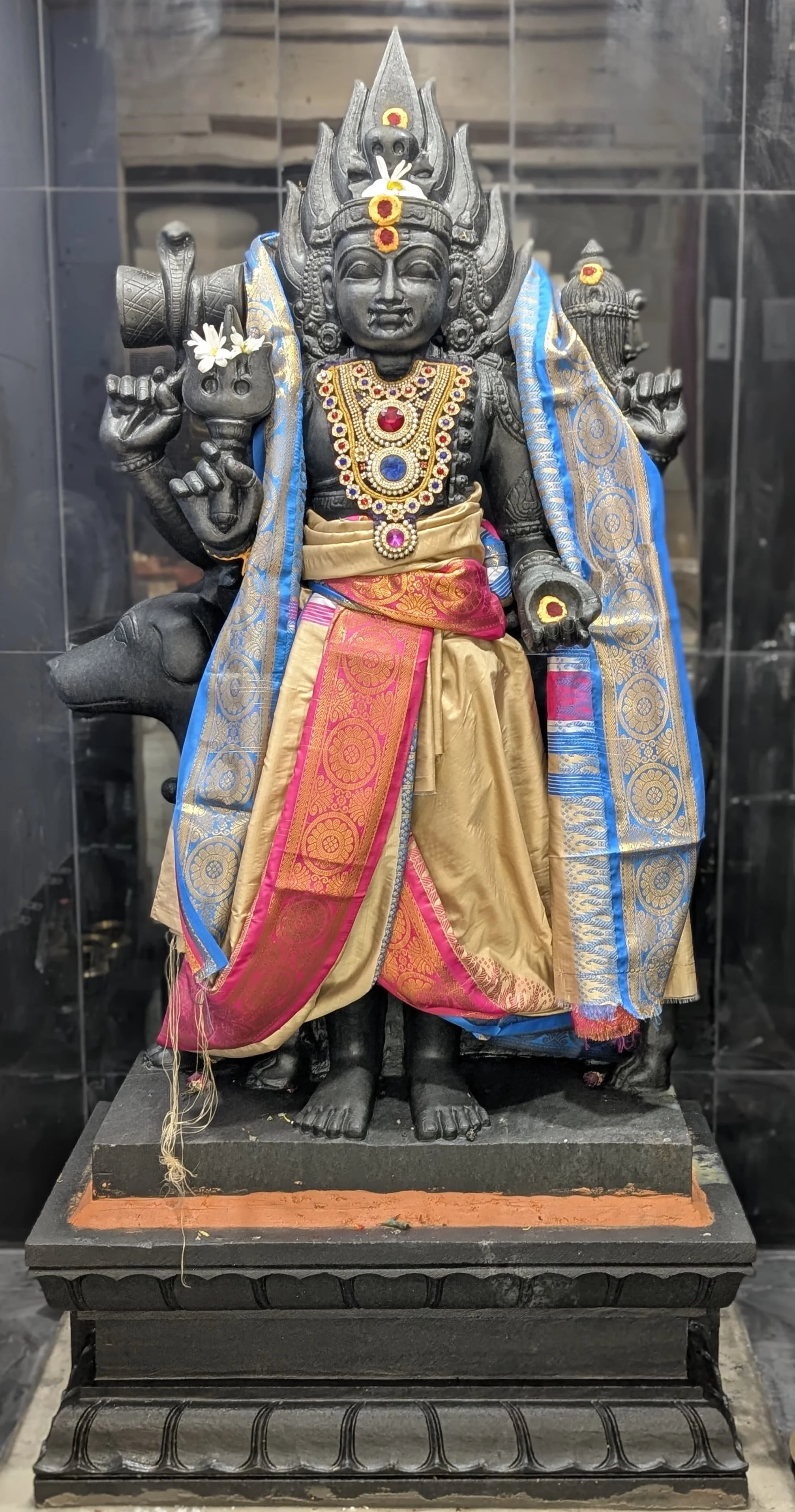 A black stone statue of a Hindu deity adorned with colorful silk garments, jewelry, and flower garlands, placed inside a glass enclosure.