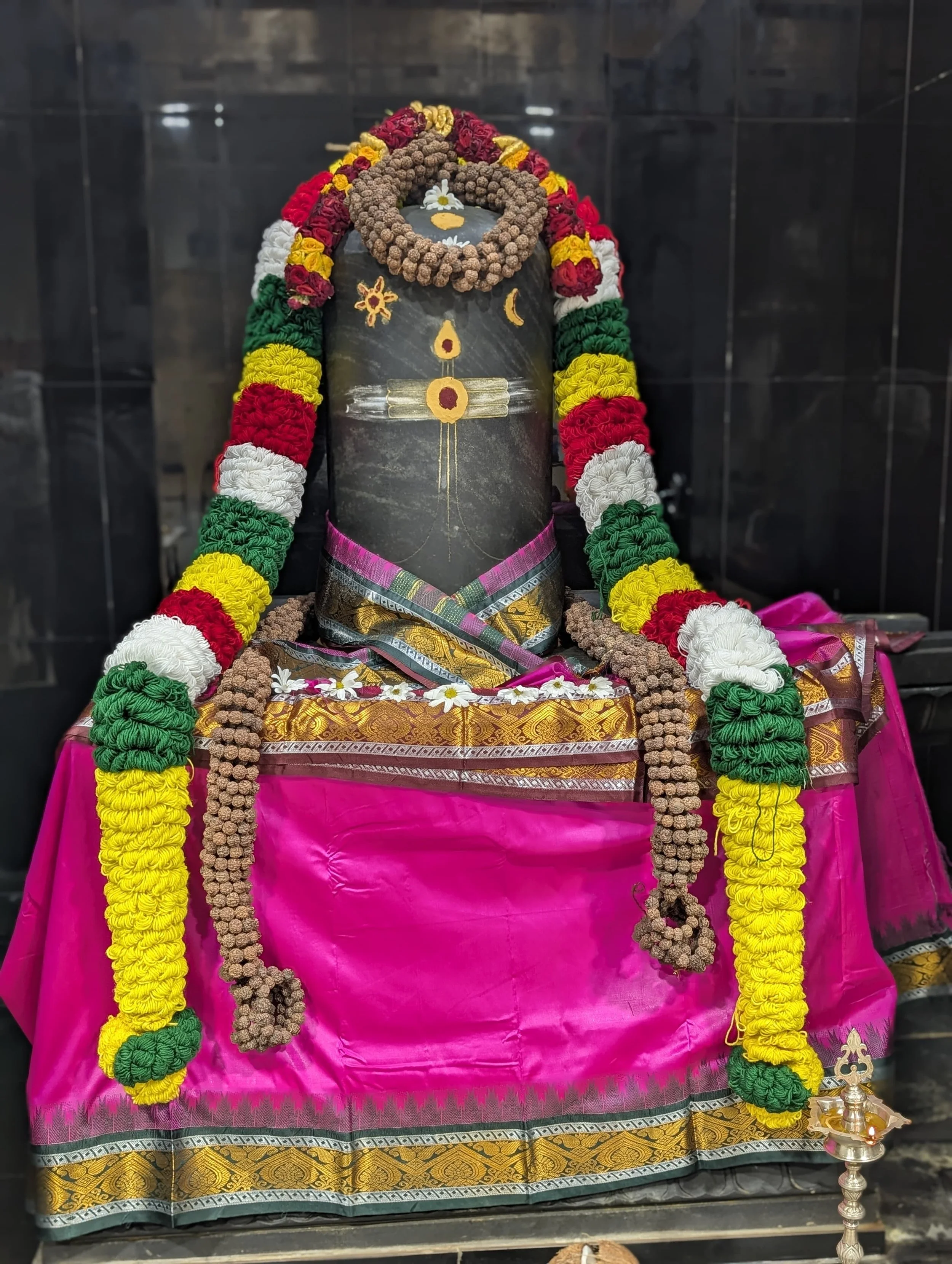A black stone idol decorated with colorful flower garlands, jewelry, and cloth, set on a pink cloth-covered platform with a black background.
