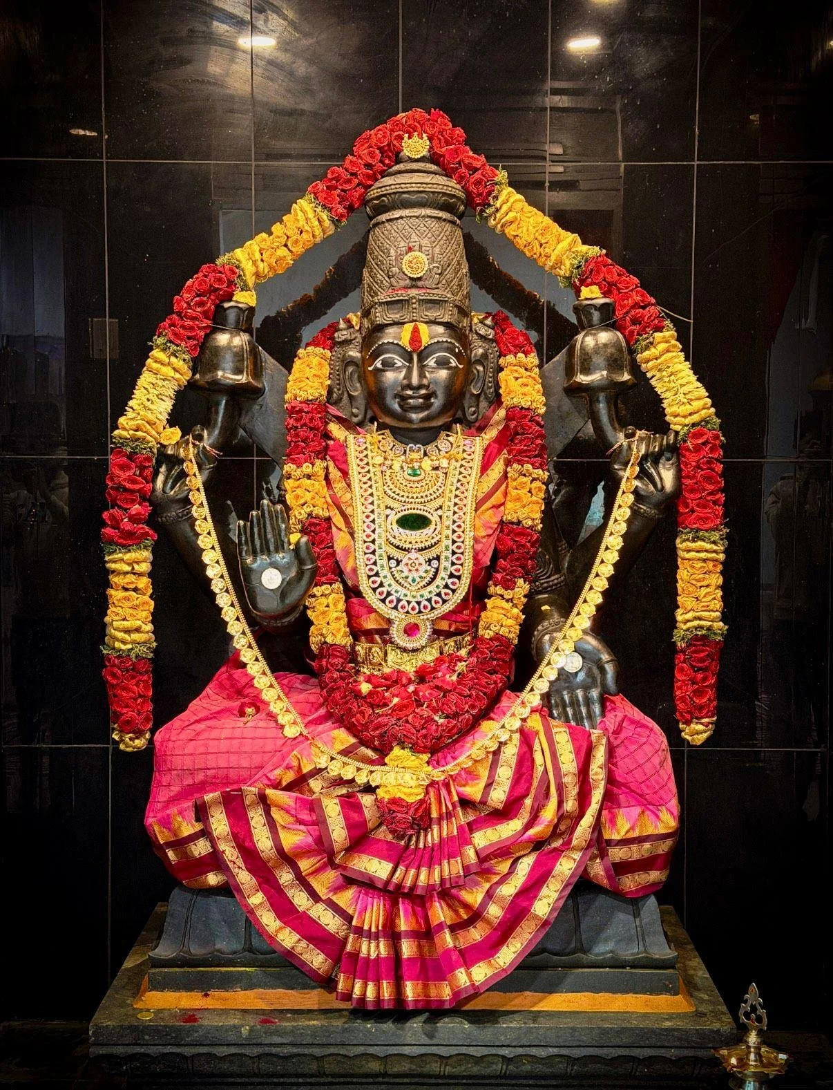 A black stone statue of a Hindu deity adorned with gold jewelry and a colorful pink and gold saree, sitting on a pedestal.
