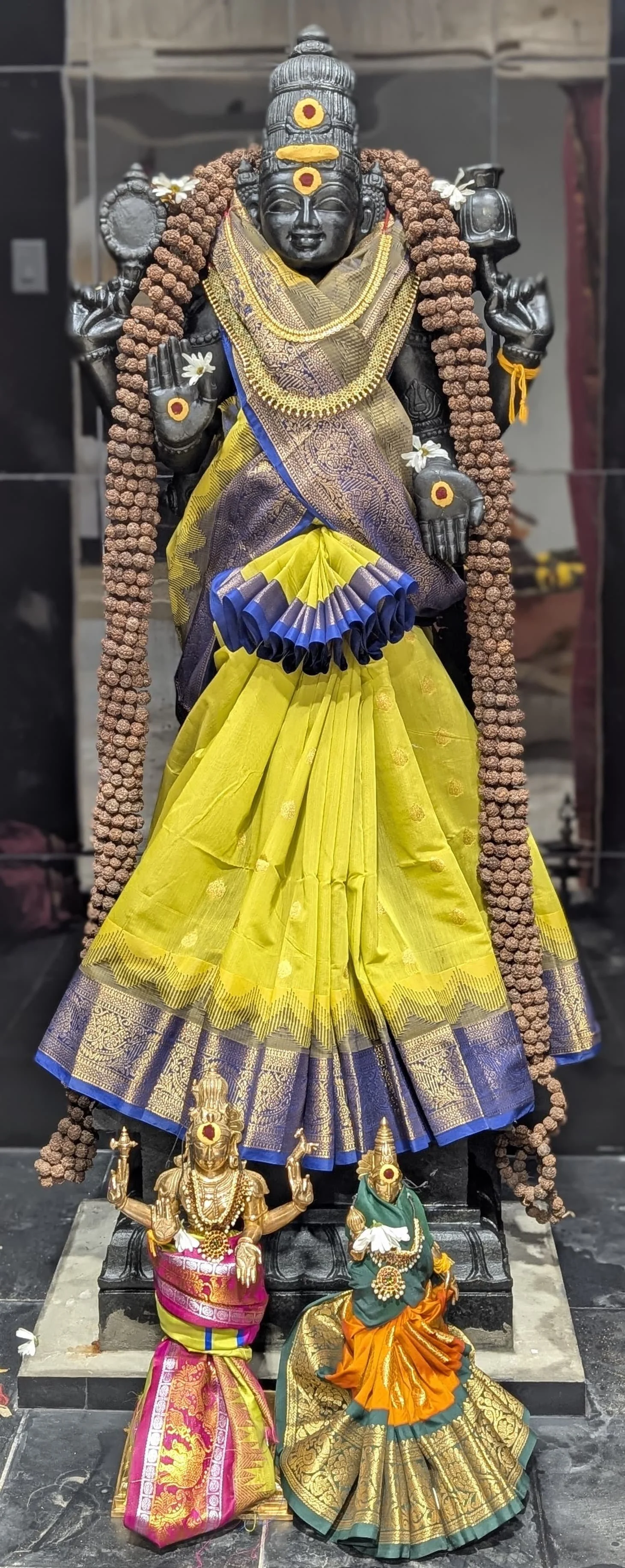Decorative Hindu deity sculpture dressed in yellow and blue silk saree, adorned with jewelry, flower garlands, and traditional ornaments, with two smaller deity statues in colorful silk garments at the base.
