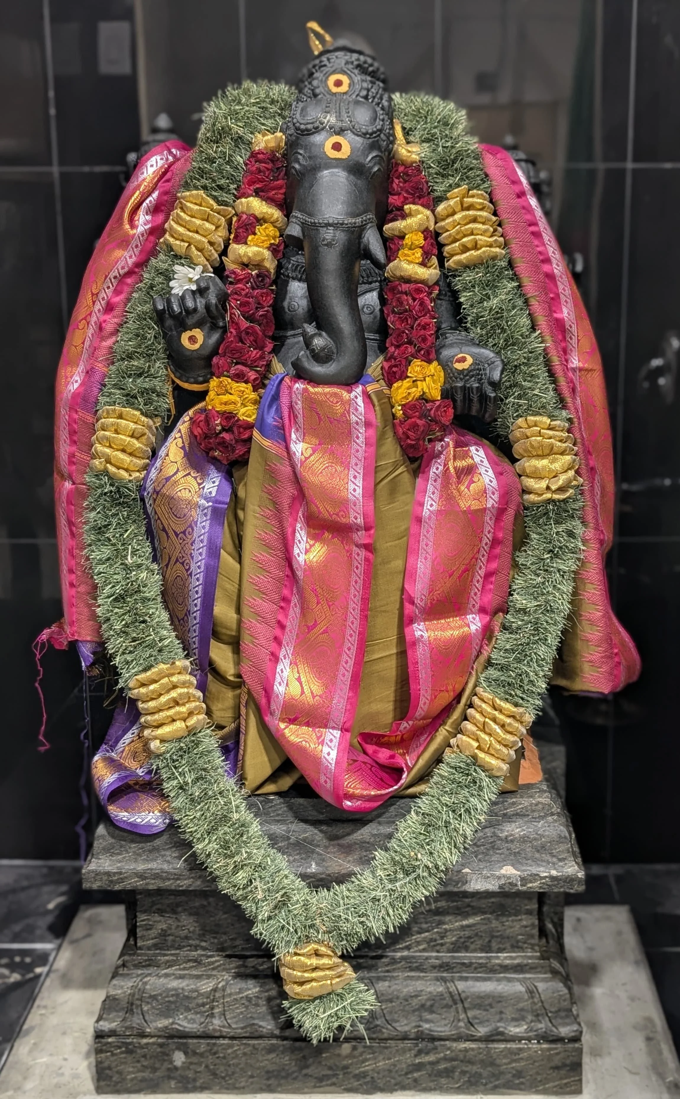 A statue of Lord Ganesha decorated with a colorful silk cloth, garlands of flowers, and a green and gold ornamental necklace, placed on a stone pedestal.