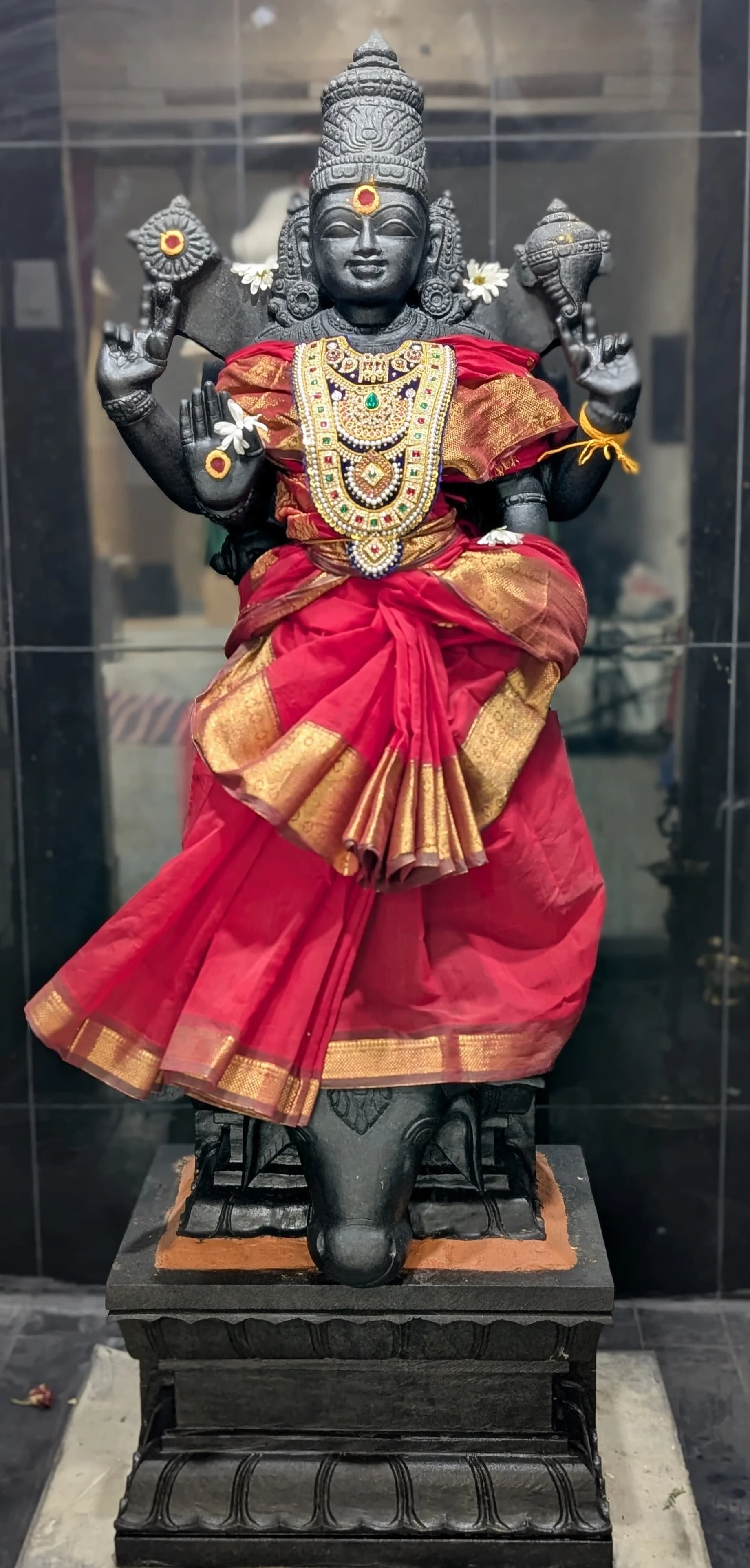Black stone sculpture of a Hindu goddess adorned with gold and jewelry, dressed in a red and gold garment, with hands in symbolic gestures, and decorated with flowers.