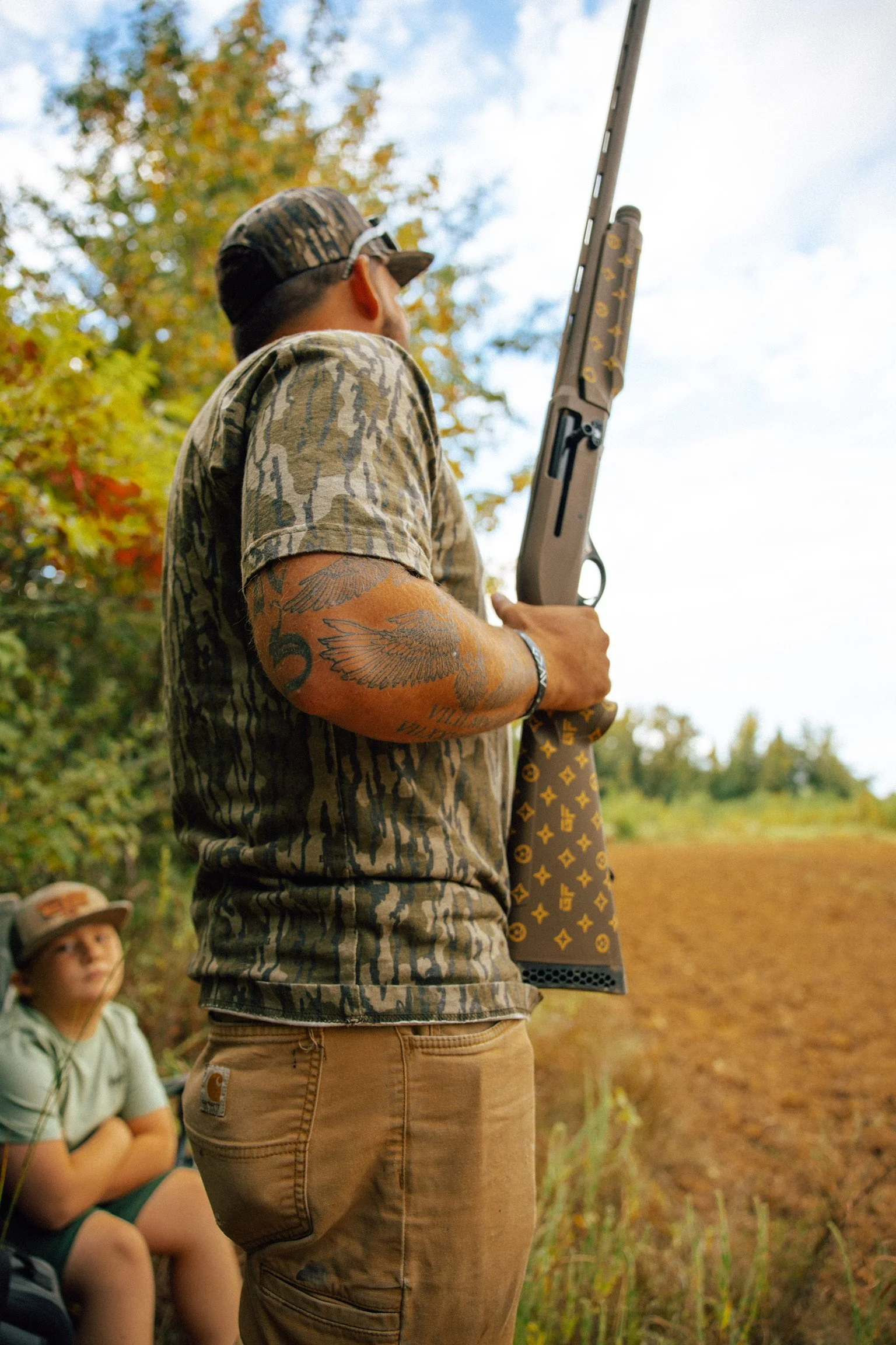 A man with camouflage clothing and a tattoo on his arm holding a rifle outdoors, with a boy sitting in the background near trees and a dirt path.