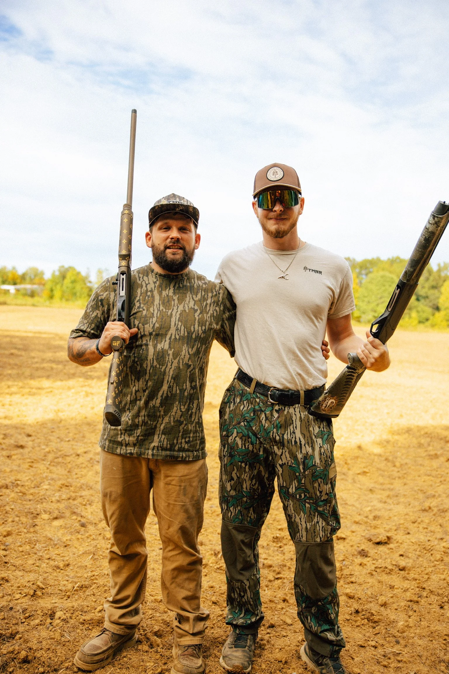 Two men standing outdoors on a dirt field, holding shotguns, dressed in camouflage or earthy tones, smiling at the camera.