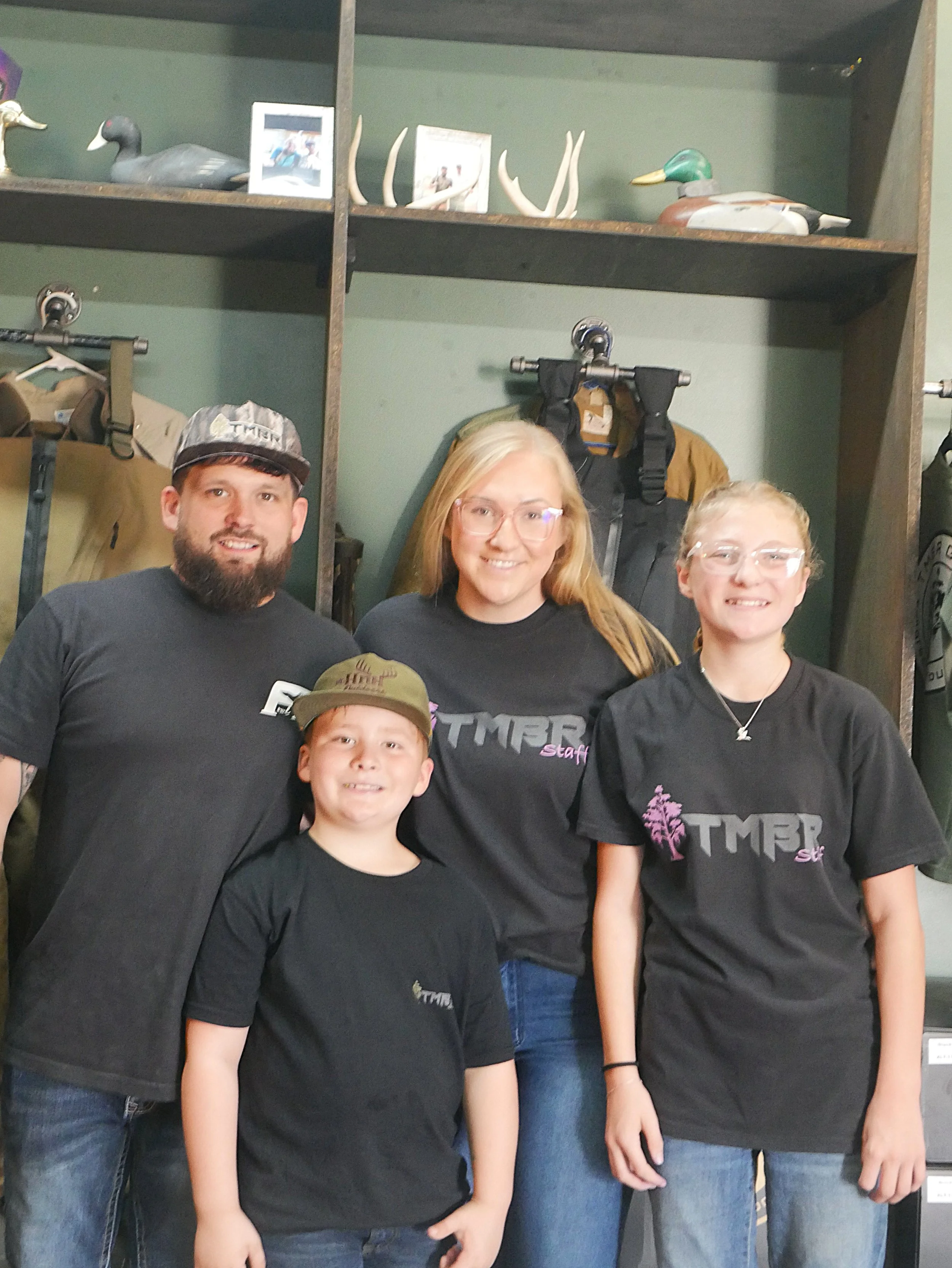 A group of four smiling people, two adults and two children, standing together in front of a wooden shelf with decorative duck and antler decorations, clothing, and bags. They are all wearing black shirts with the logo 'TMBR Staff'.