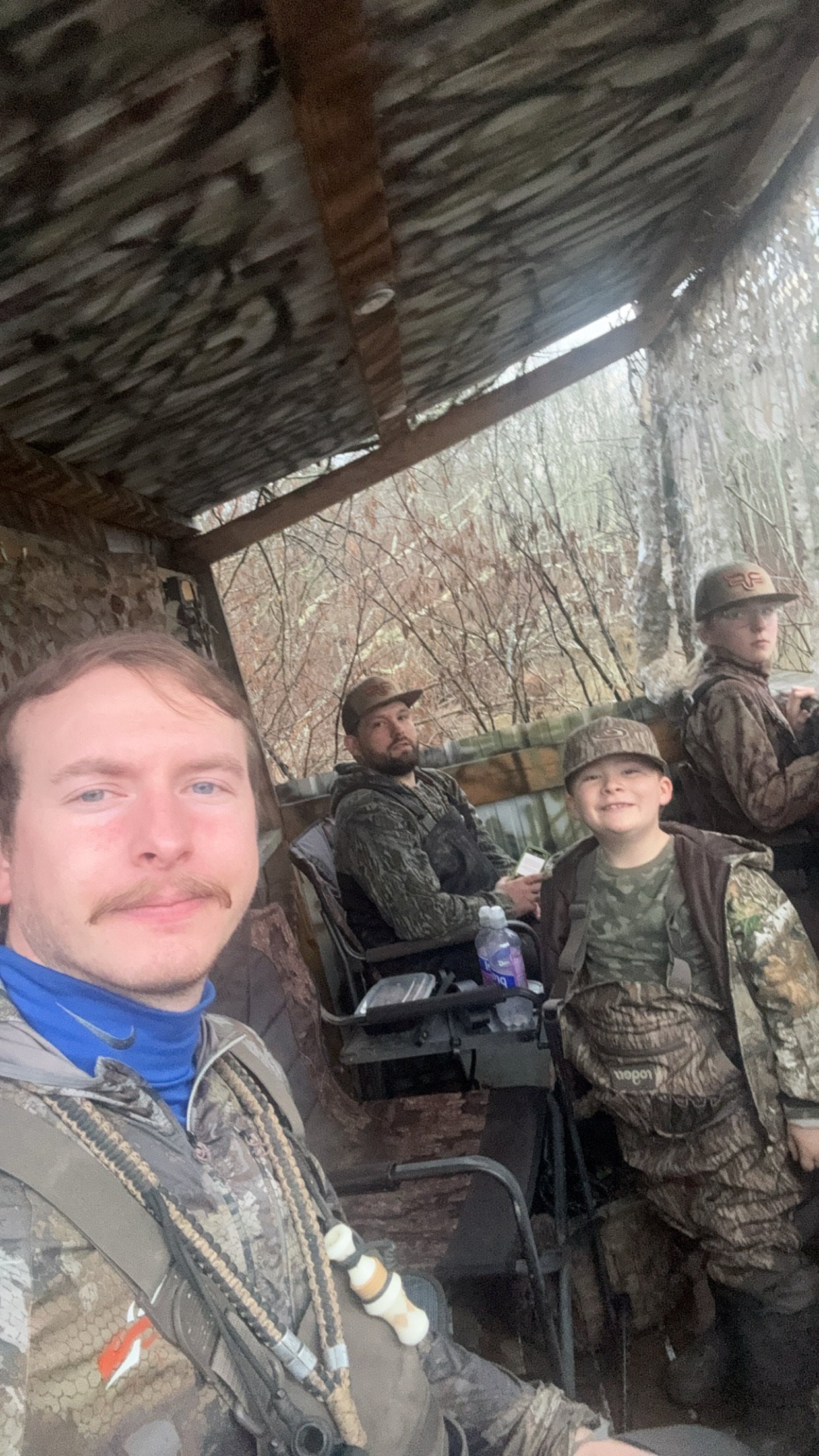 Four people sitting on outdoor chairs under a rustic wooden shelter, dressed in camouflage outdoor gear, surrounded by trees.