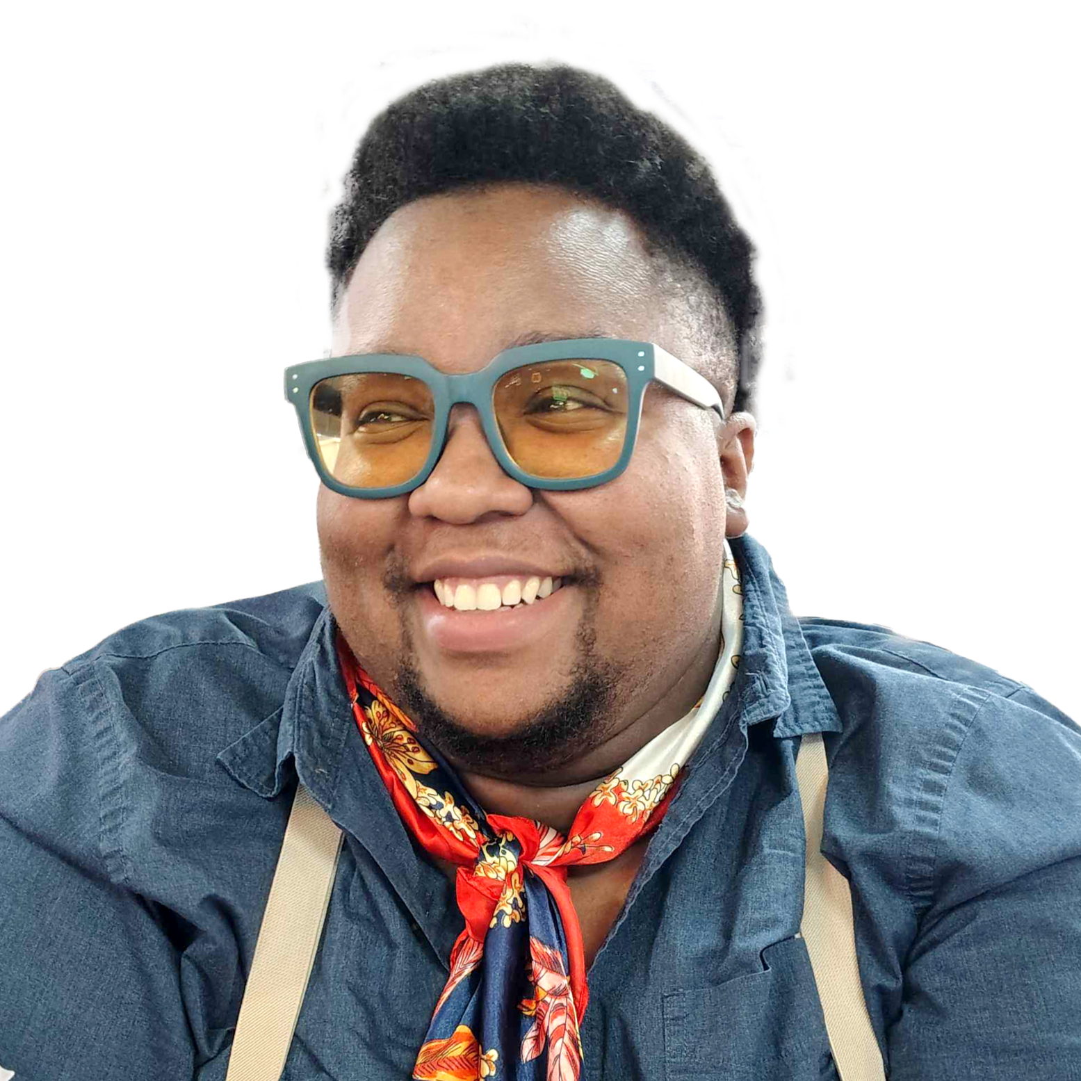 A smiling person wearing glasses with orange tinted lenses, a denim shirt, a colorful bandana around their neck, and suspenders.