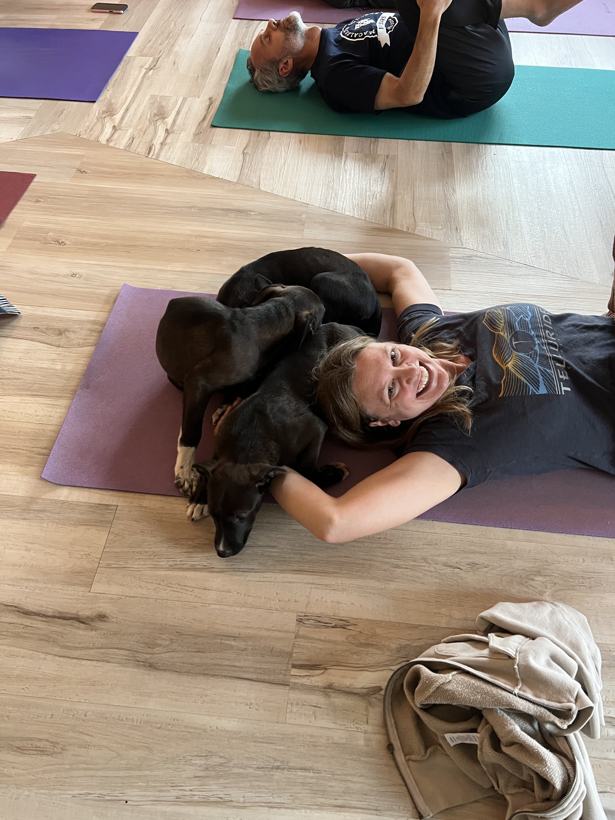 A woman lying on her back on a yoga mat with three puppies resting on her, all smiling. A man is lying on a yoga mat in the background. The setting appears to be a yoga or exercise studio with wooden floors.