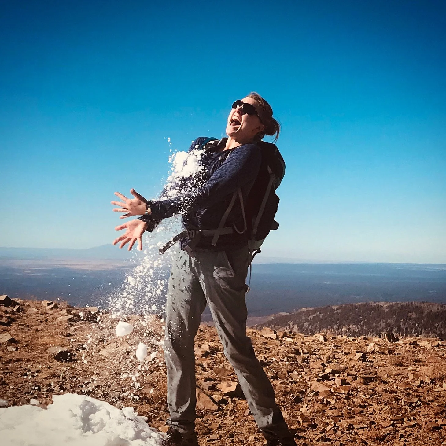 A woman with sunglasses and a backpack on a mountain top throwing snow or ice, with a clear blue sky and distant landscape in the background.