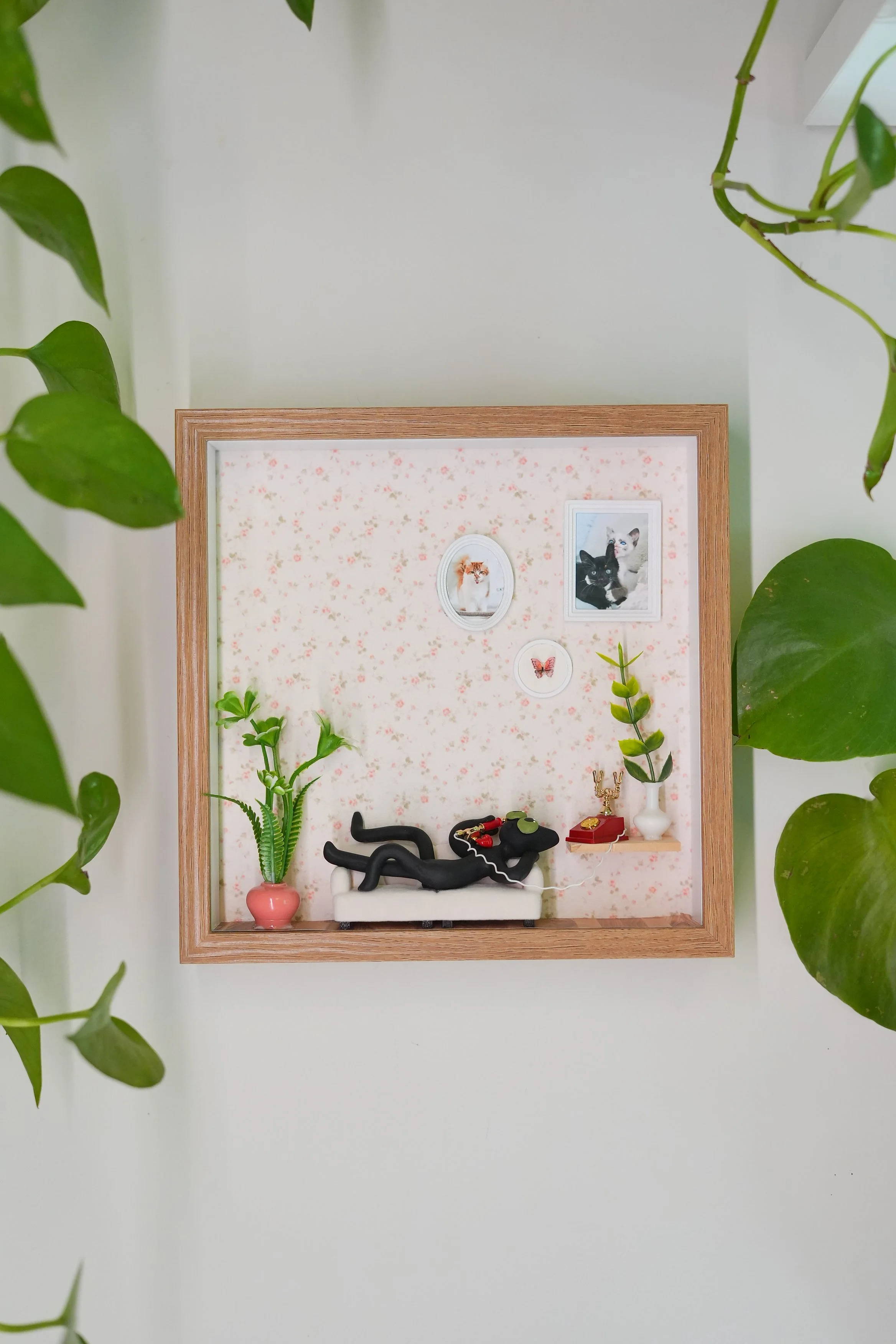 A framed collection of small decorative items and photos surrounded by green houseplants on a white wall.