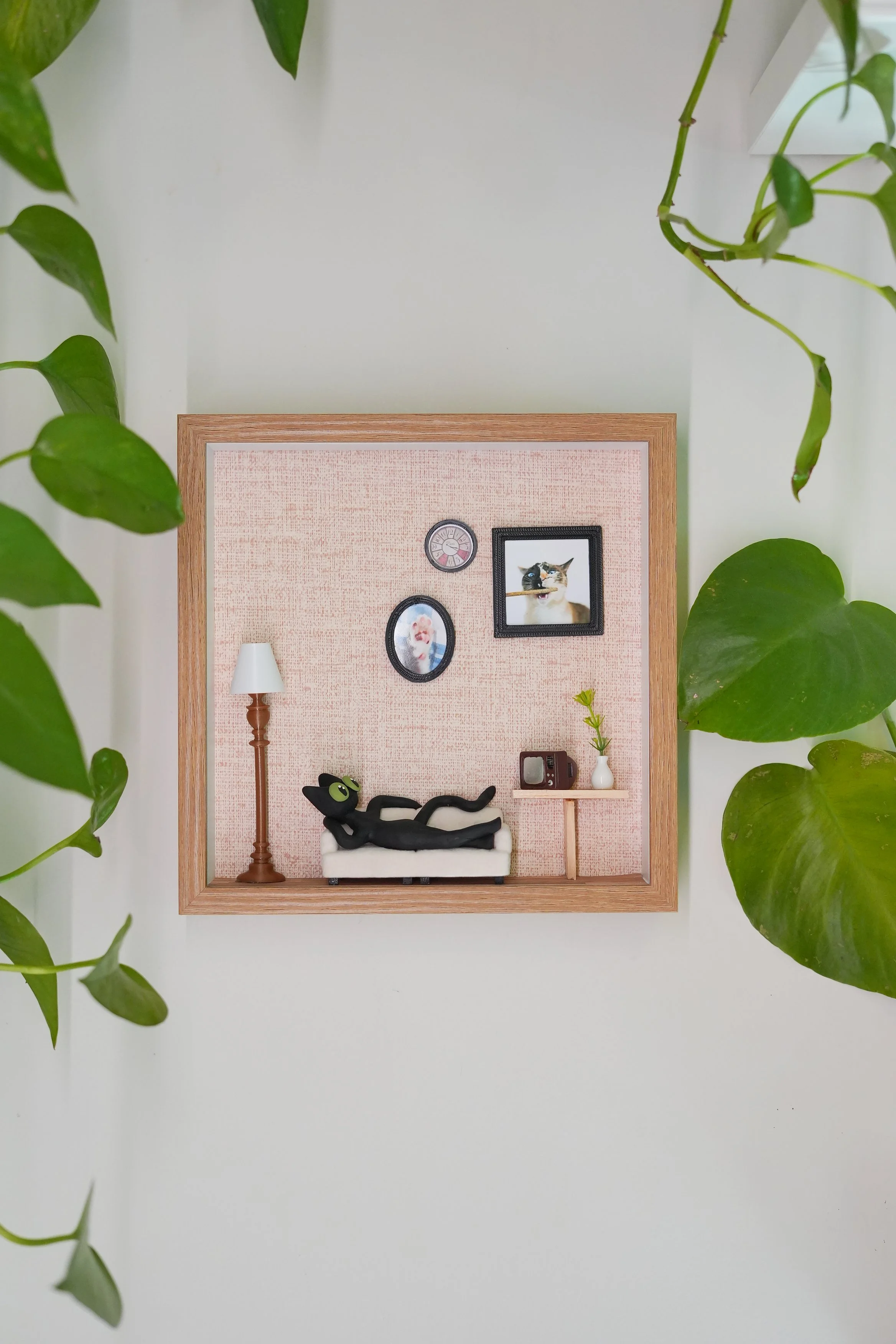 Miniature cardboard room with a black cat figurine lounging on a white couch, a small wooden side table, a white lamp, framed photos, a vintage TV, and green plant leaves framing the scene.