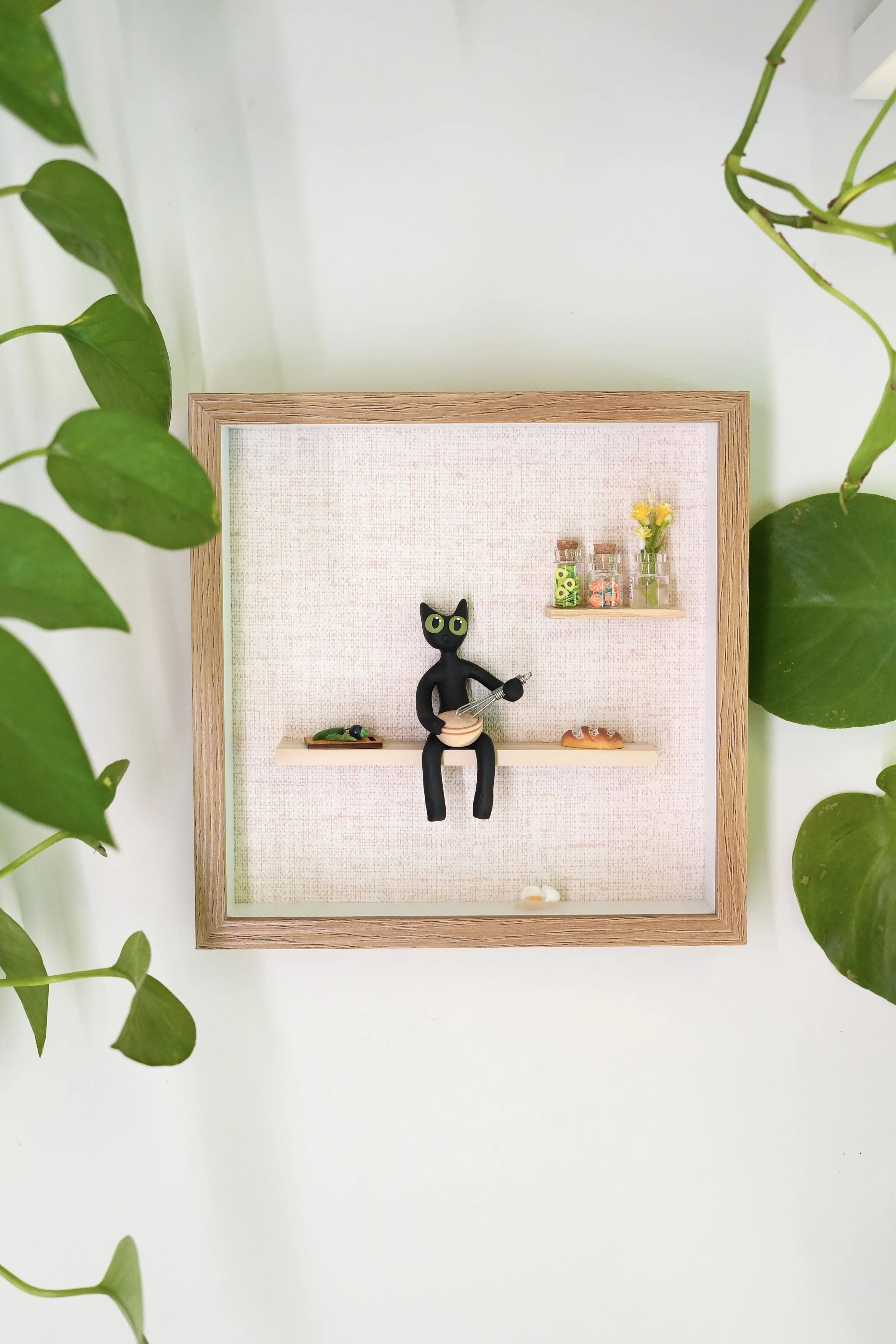 Decorative shadow box frame containing small black cat figurine, miniature glass jars with preserved flowers and greenery, and small decorative food items, surrounded by green ivy leaves.